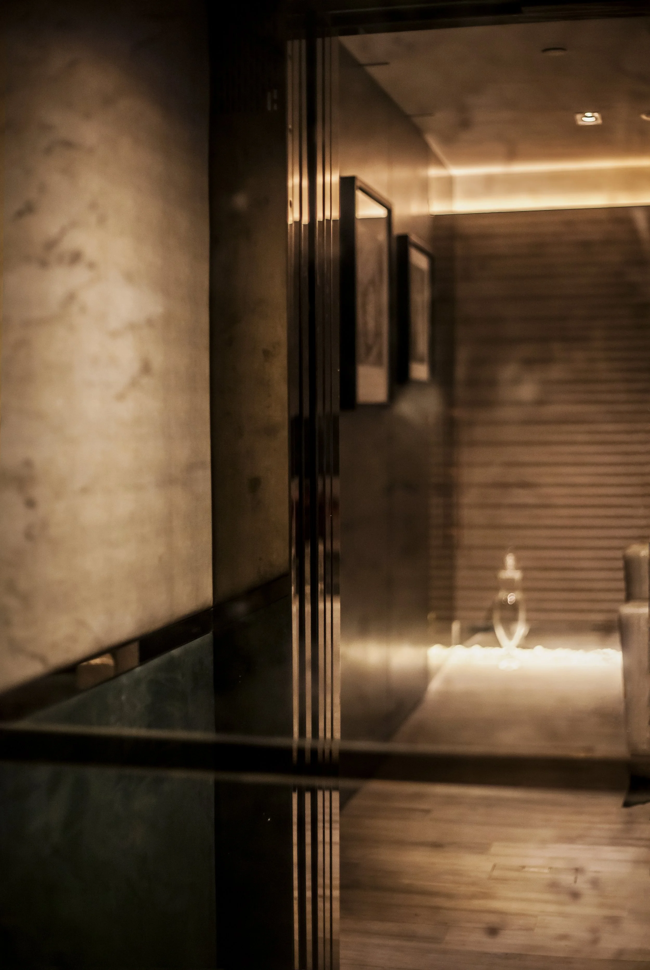 A dimly lit modern interior with wooden and metal decor, including framed artwork on the wall, a glass of water on a table, and a textured wooden wall in the background.