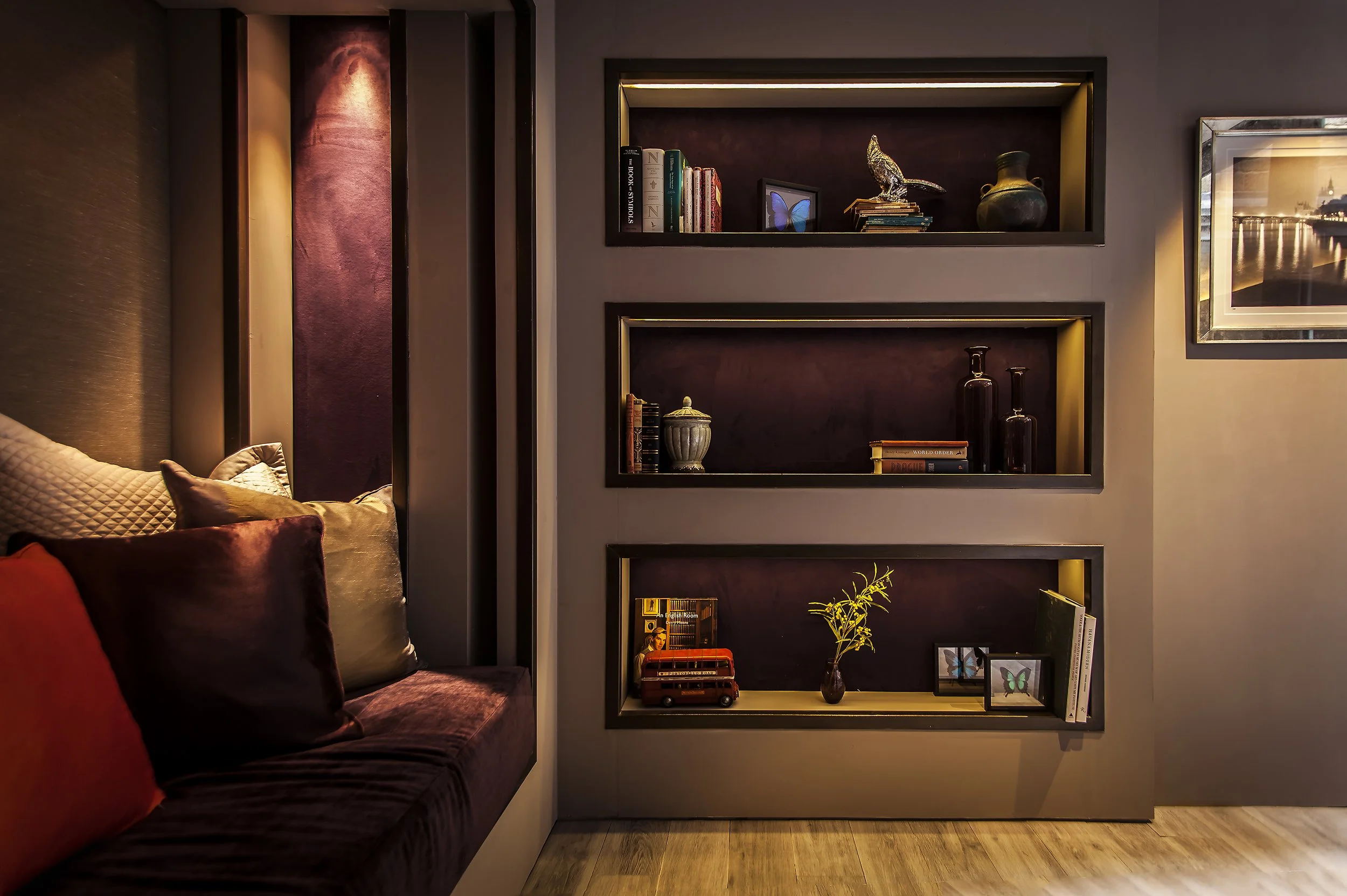 Living room with built-in dark wood shelves filled with books, decorative items, and small art pieces, next to a wall with framed artwork, and part of a cozy seating area with pillows.