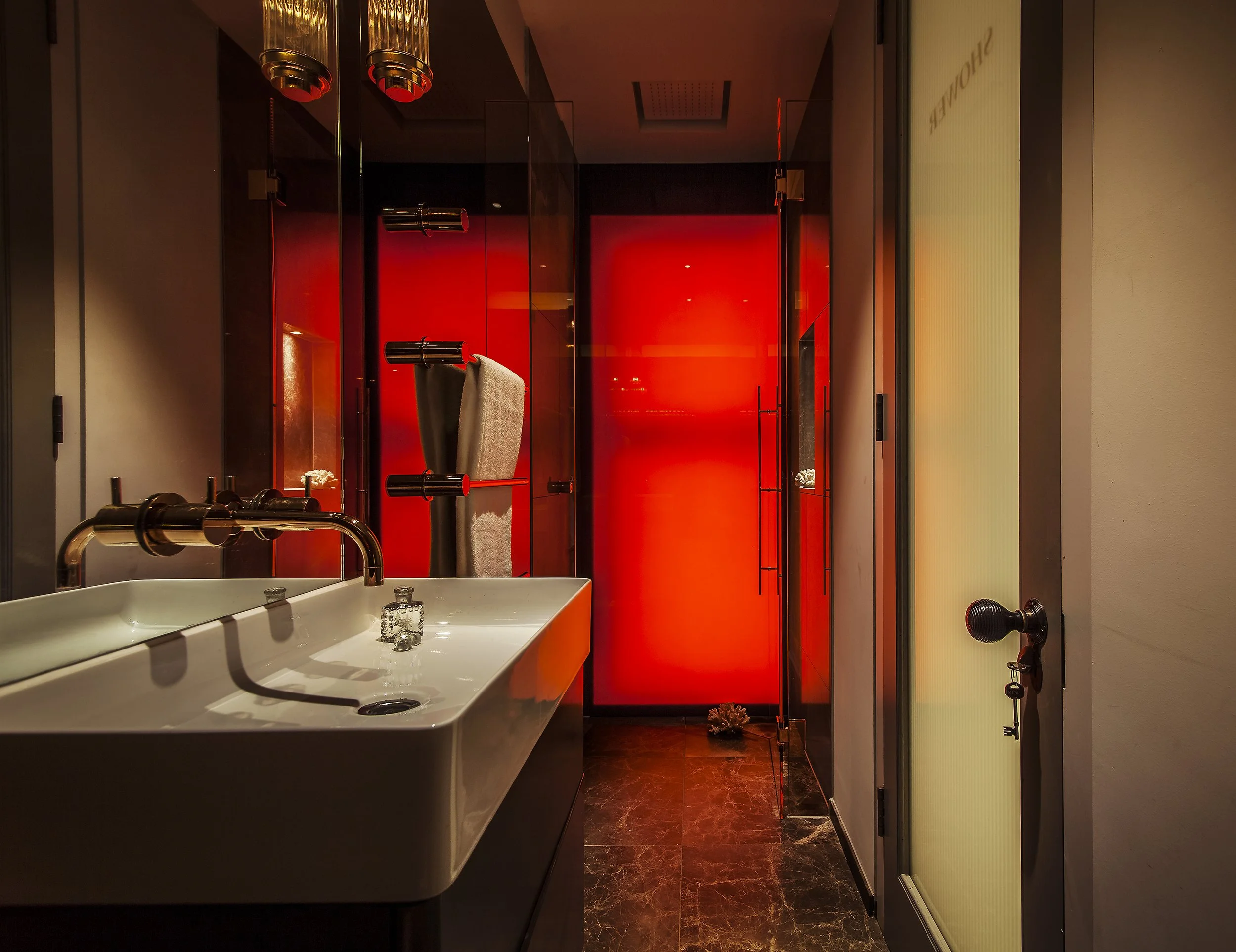 Modern bathroom with a white sink, chrome faucet, beige towel on a towel warmer, and a red-lit shower or changing area at the back.