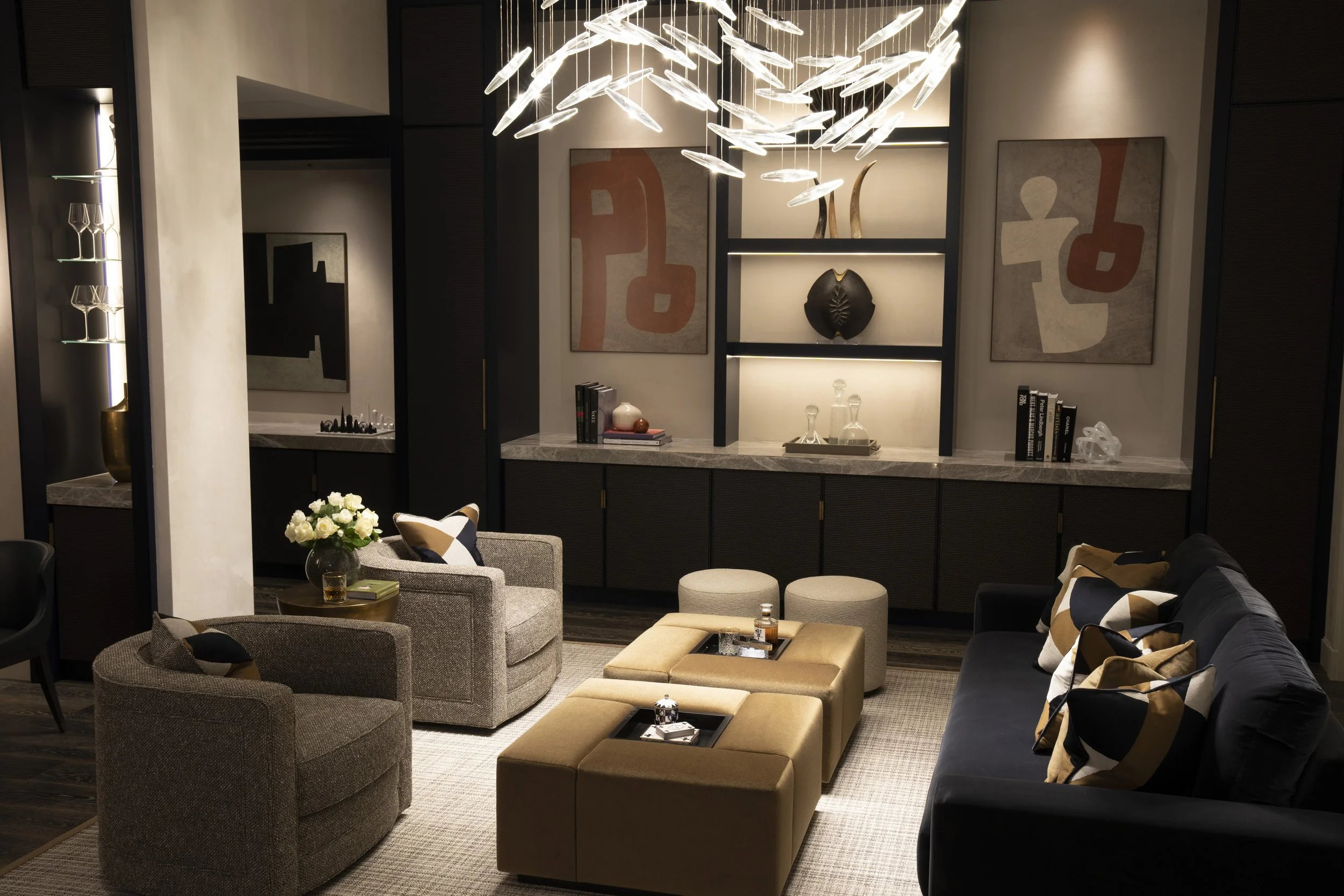 Elegant modern living room with a black sofa, two beige armchairs, and a set of beige ottomans, decorated with stylish throw pillows. A marble-topped cabinet with decorative items and books is against the back wall, featuring abstract art pieces on t