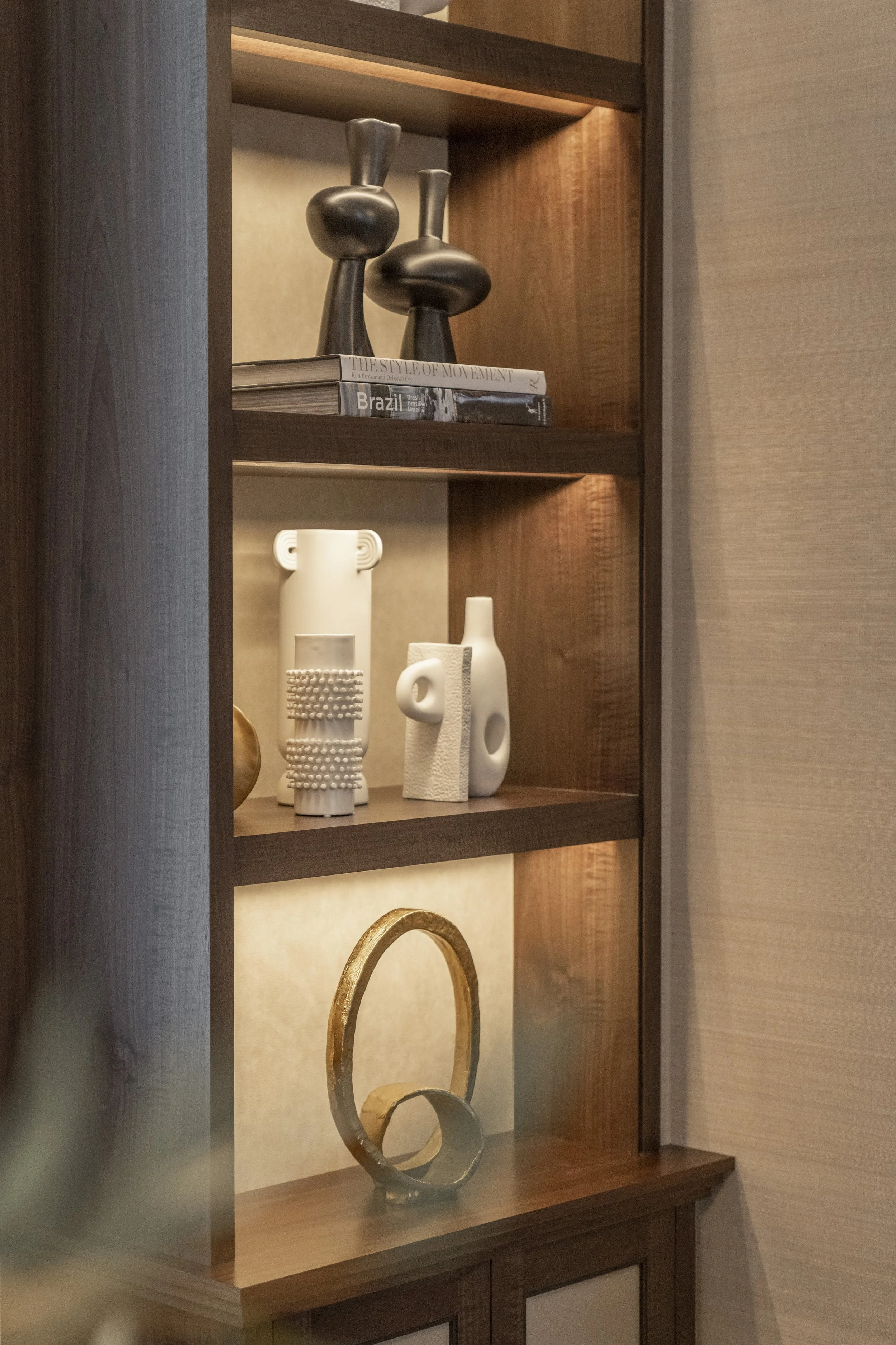 A wooden display cabinet with three shelves holding decorative objects including vases, books, and sculptures in neutral tones and metallic accents.