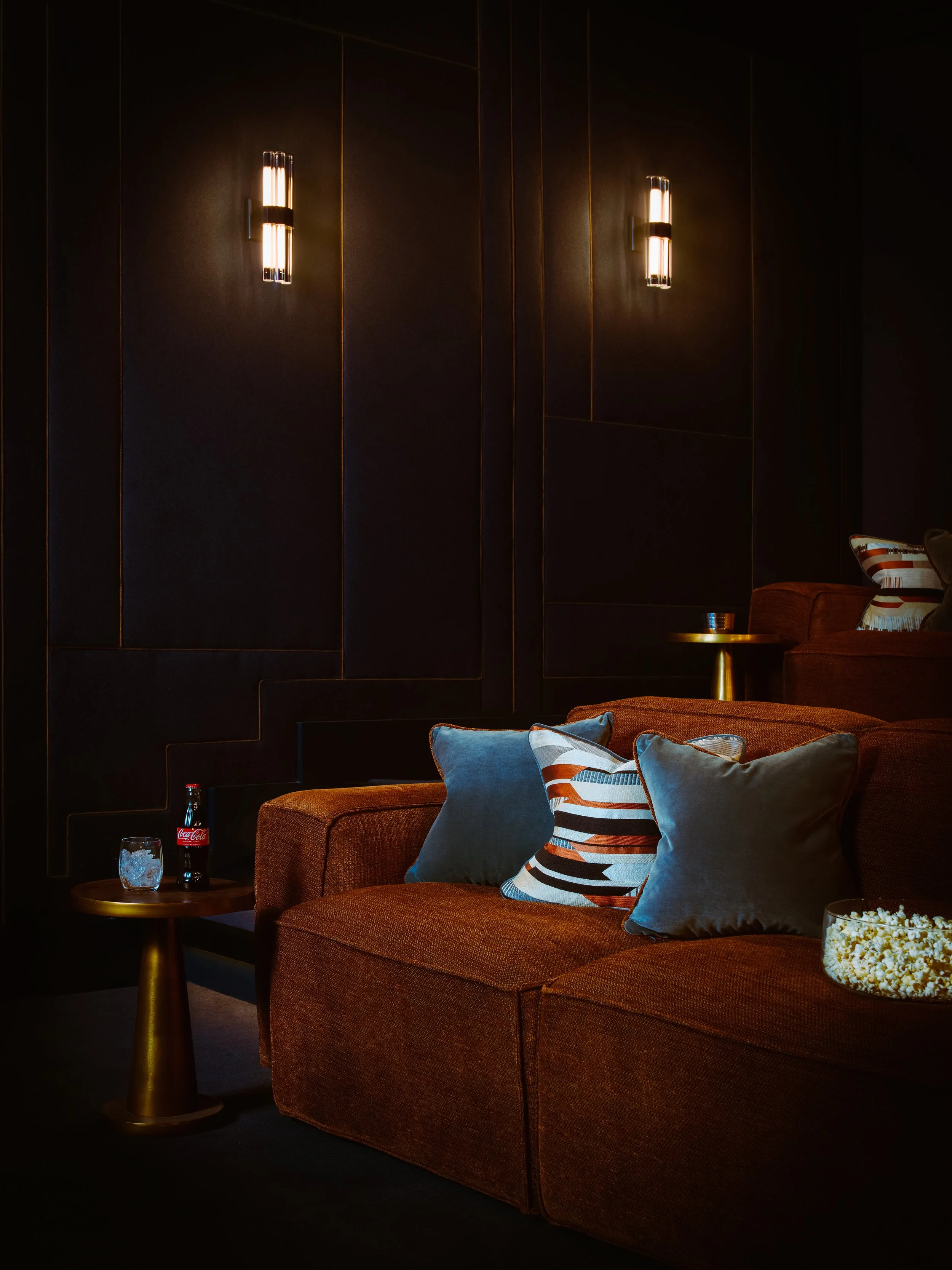 A cozy living room corner with a brown sofa, decorative pillows, a small side table with a glass of ice, a Coca-Cola bottle, and a bowl of popcorn, against dark paneled walls with modern wall-mounted lights.