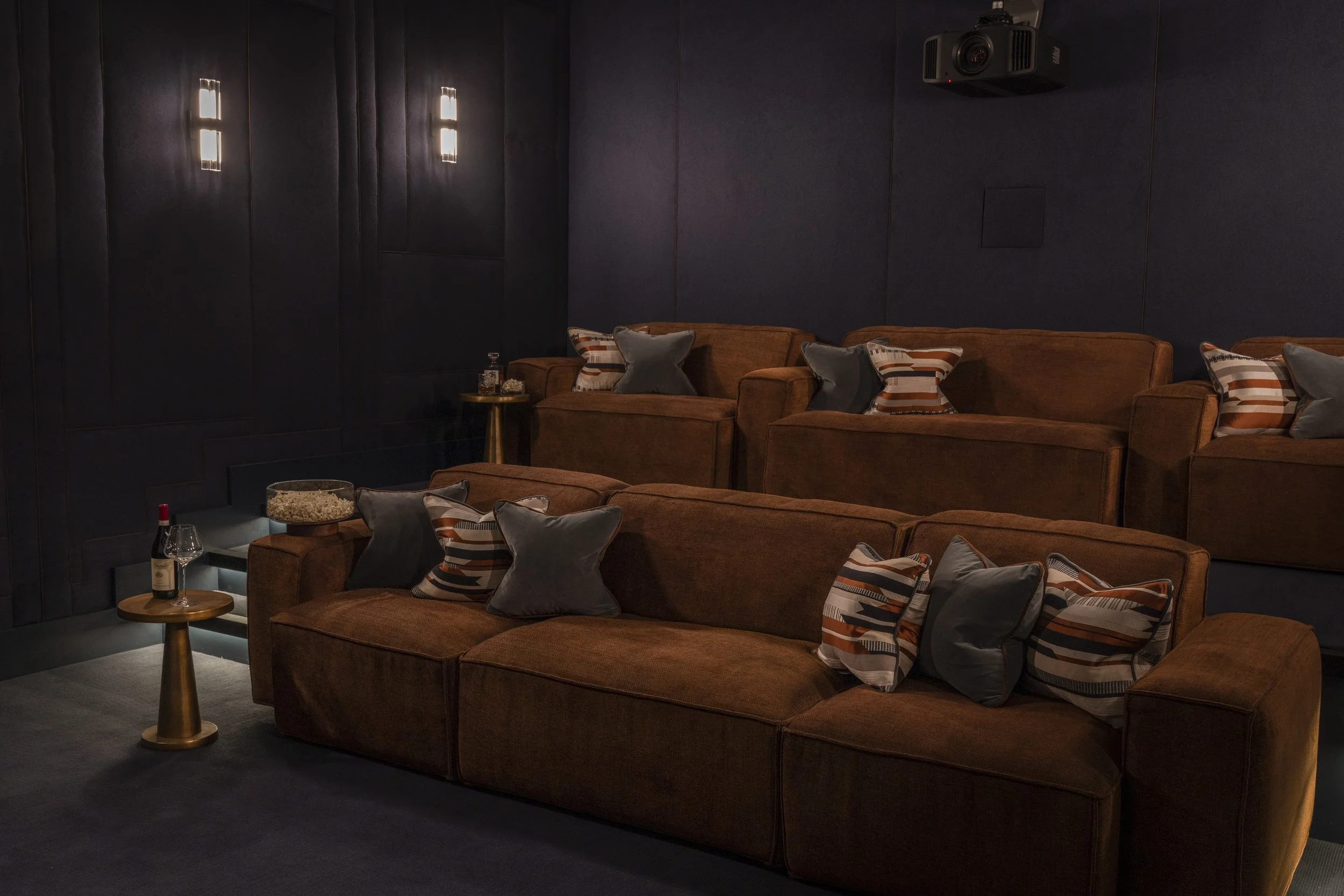 A dimly lit lounge with dark navy walls and six brown velvet sofas. Each sofa has decorative pillows in various patterns and shades of gray, brown, and striped designs. There are small round side tables with drinks and snacks, with one table holding 