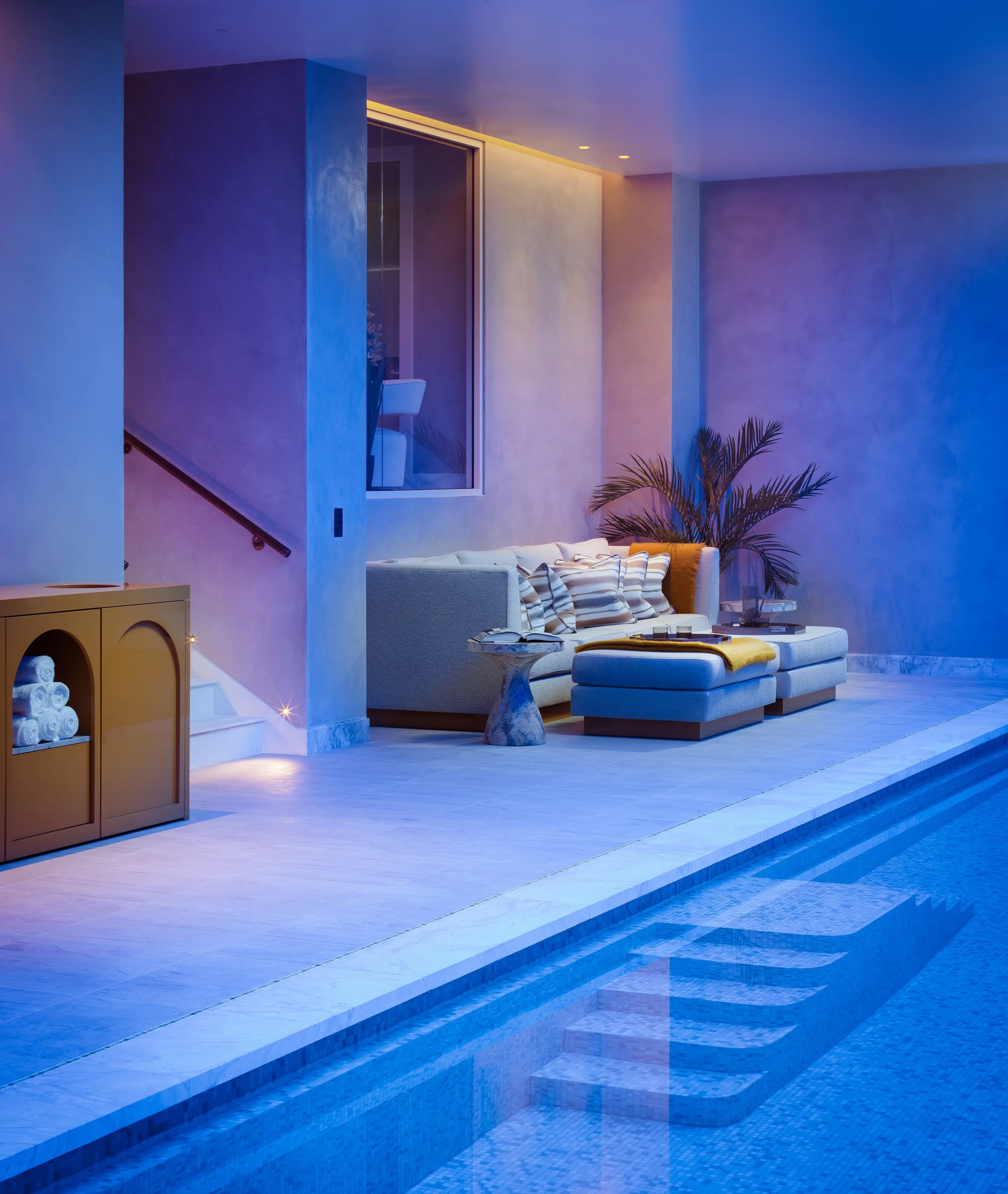 Indoor poolside seating area with a white sofa, striped cushions, small marble side table, large potted palm, and a wooden cabinet with rolled towels, illuminated by soft purple and yellow lighting.
