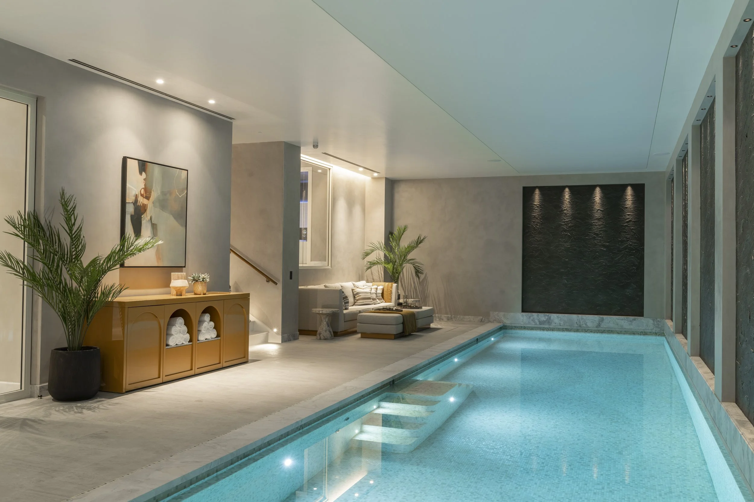 Indoor swimming pool area with a seating area, large potted plants, artwork on the wall, and ambient lighting.