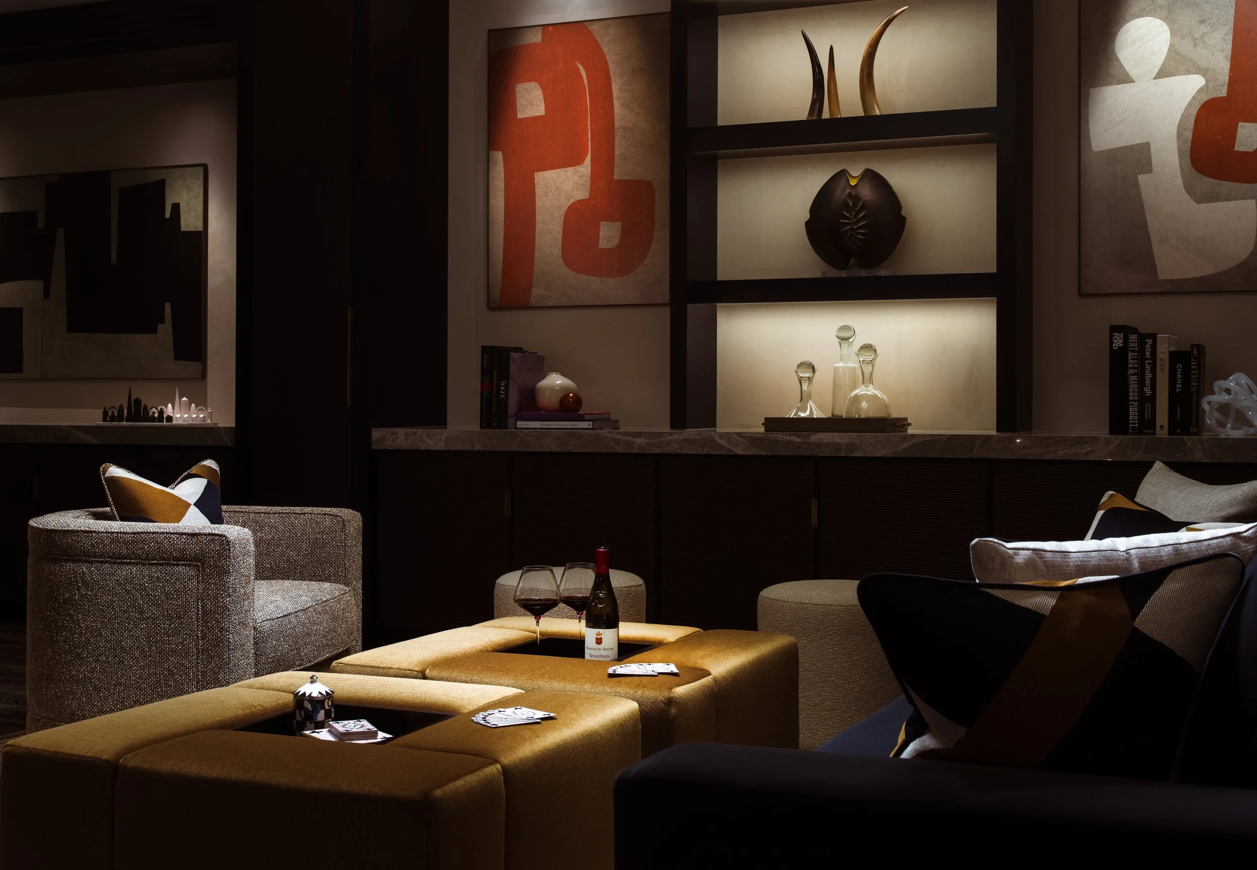 A cozy living room with modern decor, including upholstered chairs, a yellow ottoman with recessed sections holding a wine bottle and glasses, abstract artwork on the walls, and shelves with decorative items. There are also books and pillows on the f