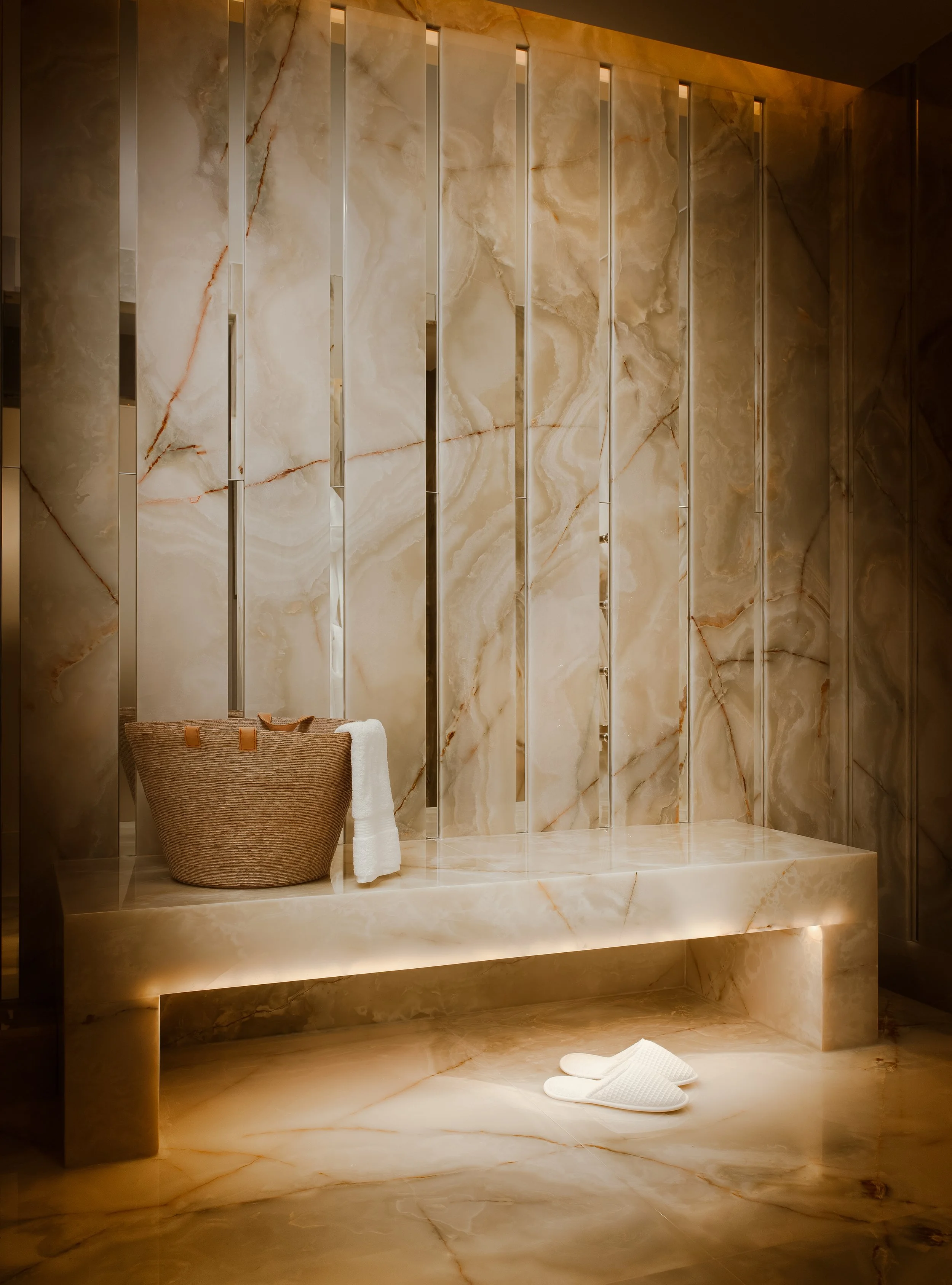 Luxurious bathroom with marble walls and floor, a woven brown basket, a white towel, and white slippers on the floor.