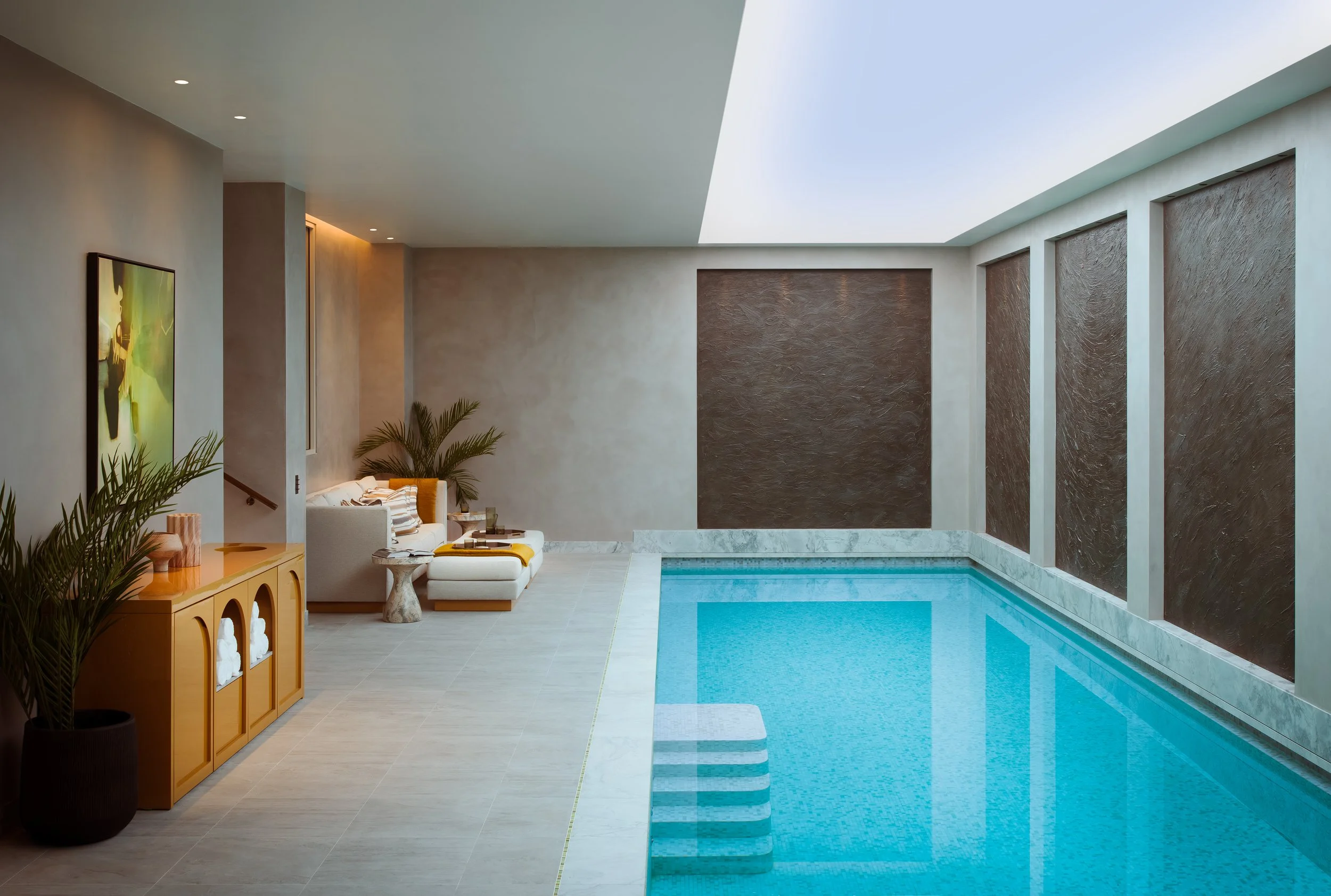 Indoor swimming pool with steps leading into the water, lounge area with white couch and pillows, potted plants, artwork on the wall, and a large skylight overhead illuminating the space.