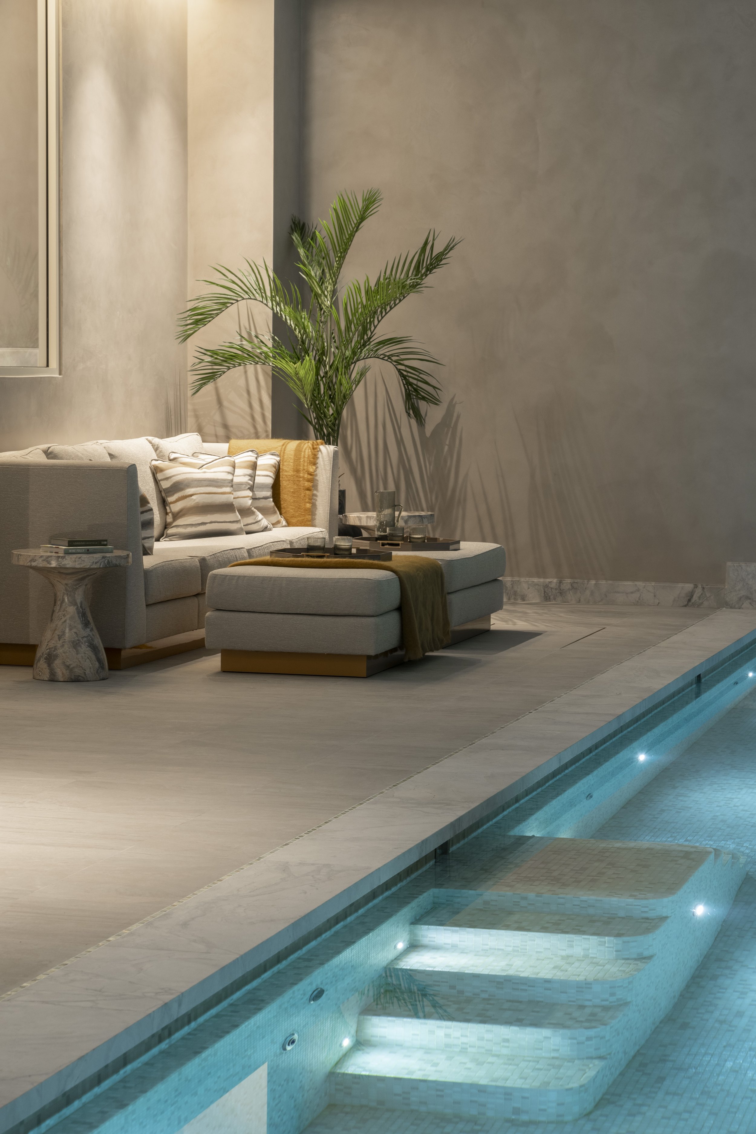 Lounge area adjacent to a swimming pool with a beige sofa, striped cushions, a green throw, a marble side table, and a tall green plant, in a modern indoor setting.
