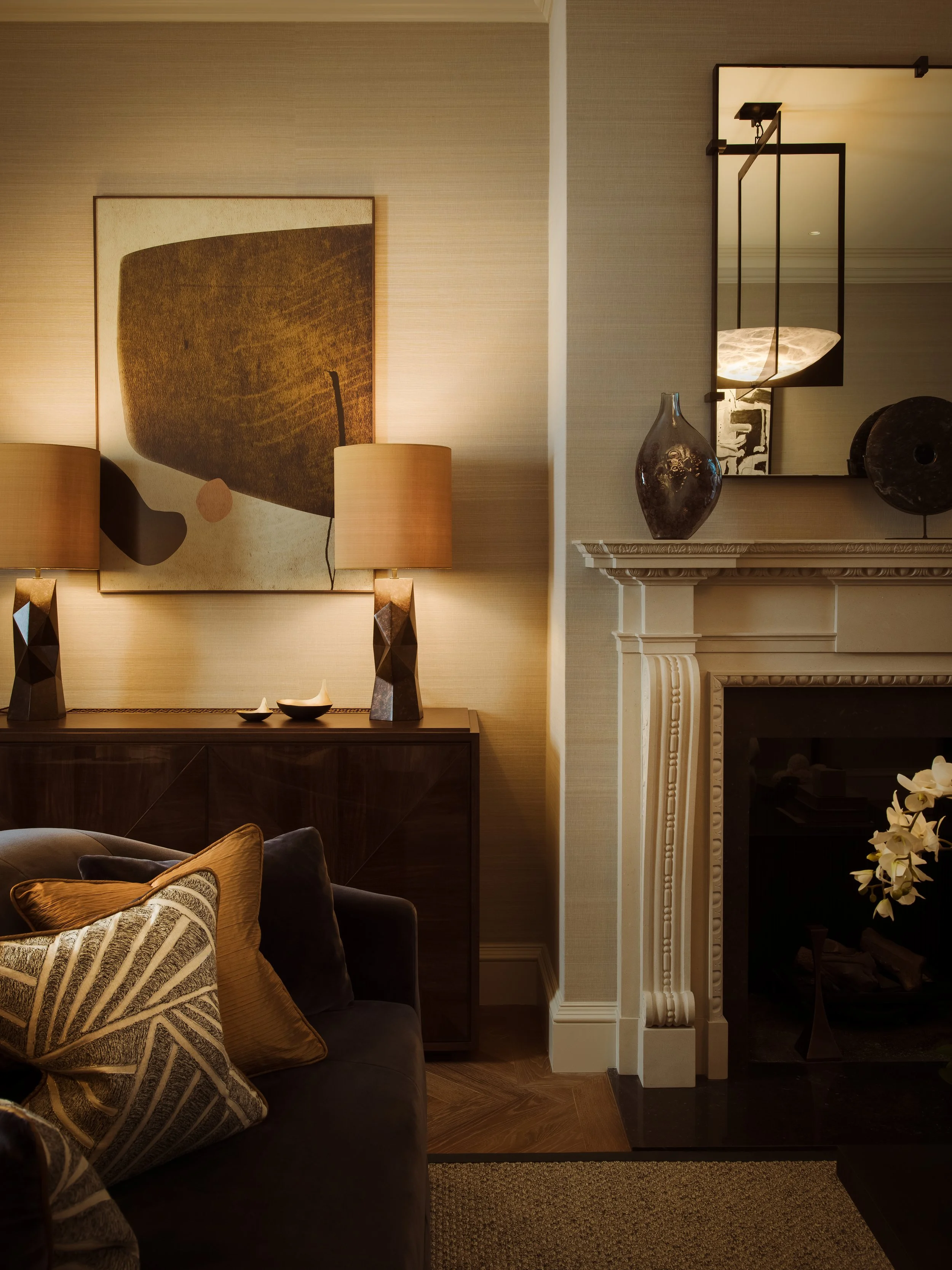 A living room corner featuring a dark sofa with decorative pillows, a wooden sideboard with two lamps and decorative bowls, modern artwork above, and a fireplace with a mirror, vase, and sculpture on the mantel.