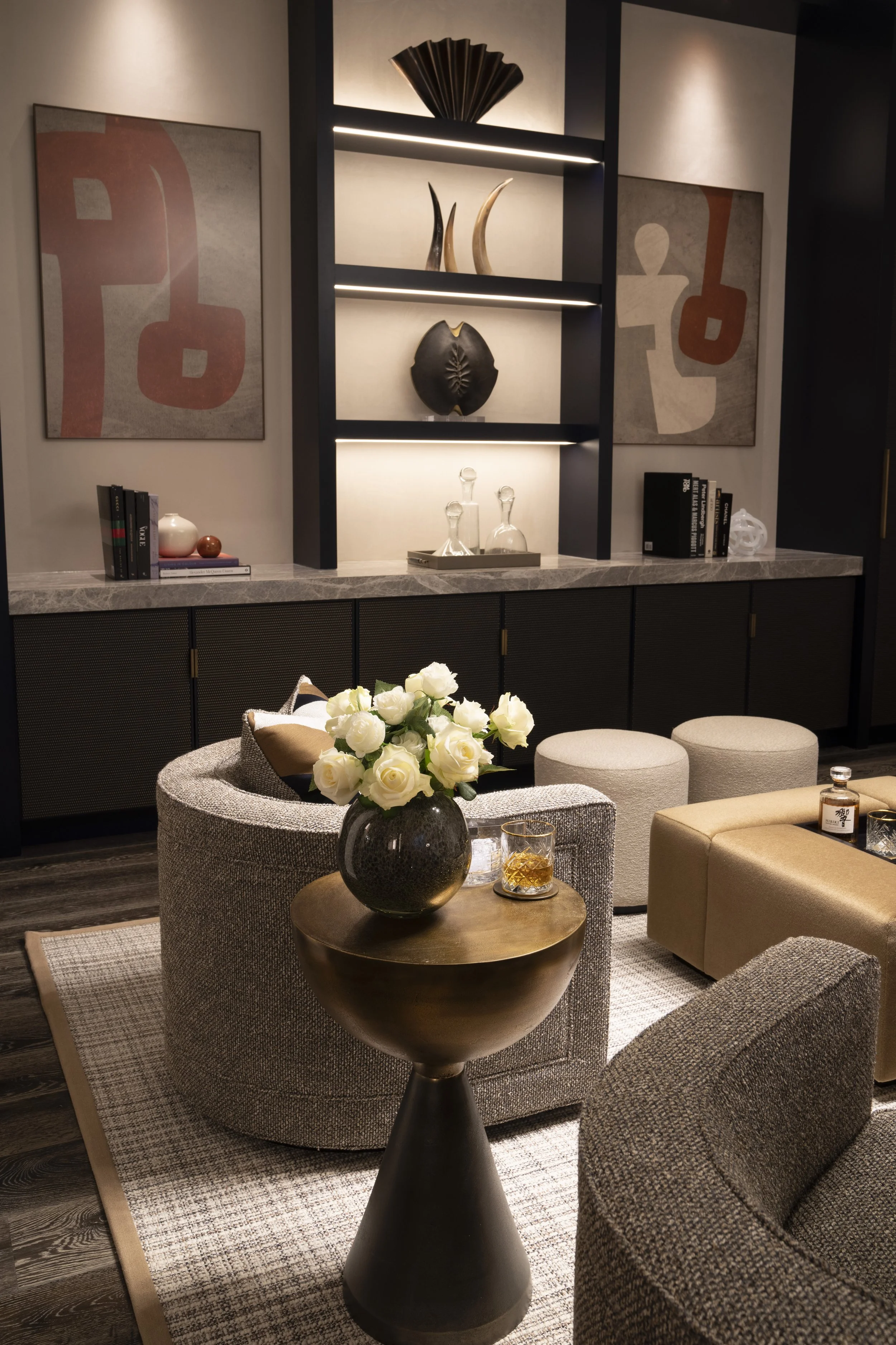 Modern living room with a round beige sofa, a black vase with white roses on a small metallic table, abstract wall art, decorative shelves with sculptures, and a marble-topped cabinet.