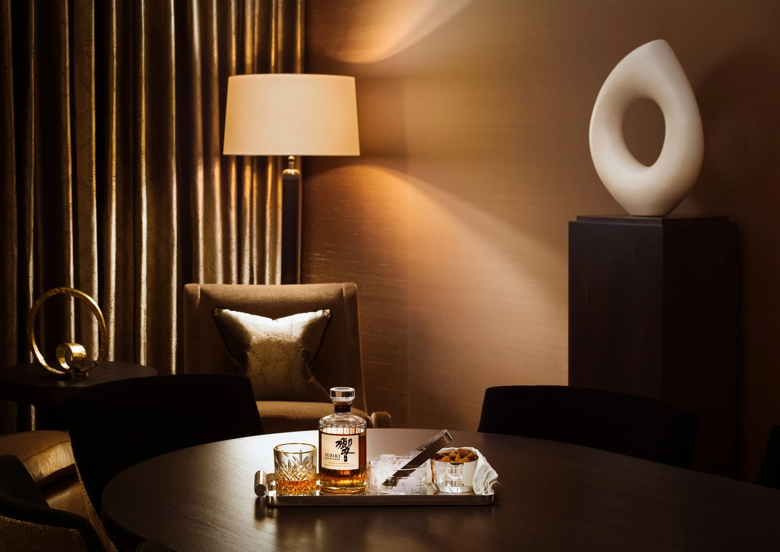 Elegant dining room with a round table holding a bottle of whiskey, a glass of whiskey, and snacks, surrounded by dark chairs, with a lamp, a gold decorative object, a cushioned armchair, curtains, and a sculptural decorative piece on a pedestal