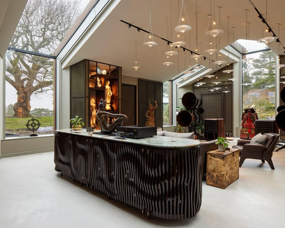 Modern interior living space with large glass windows showing a large tree outside, and a mix of contemporary and eclectic decor including sculptures, plants, and artwork, with seating and lighting fixtures.