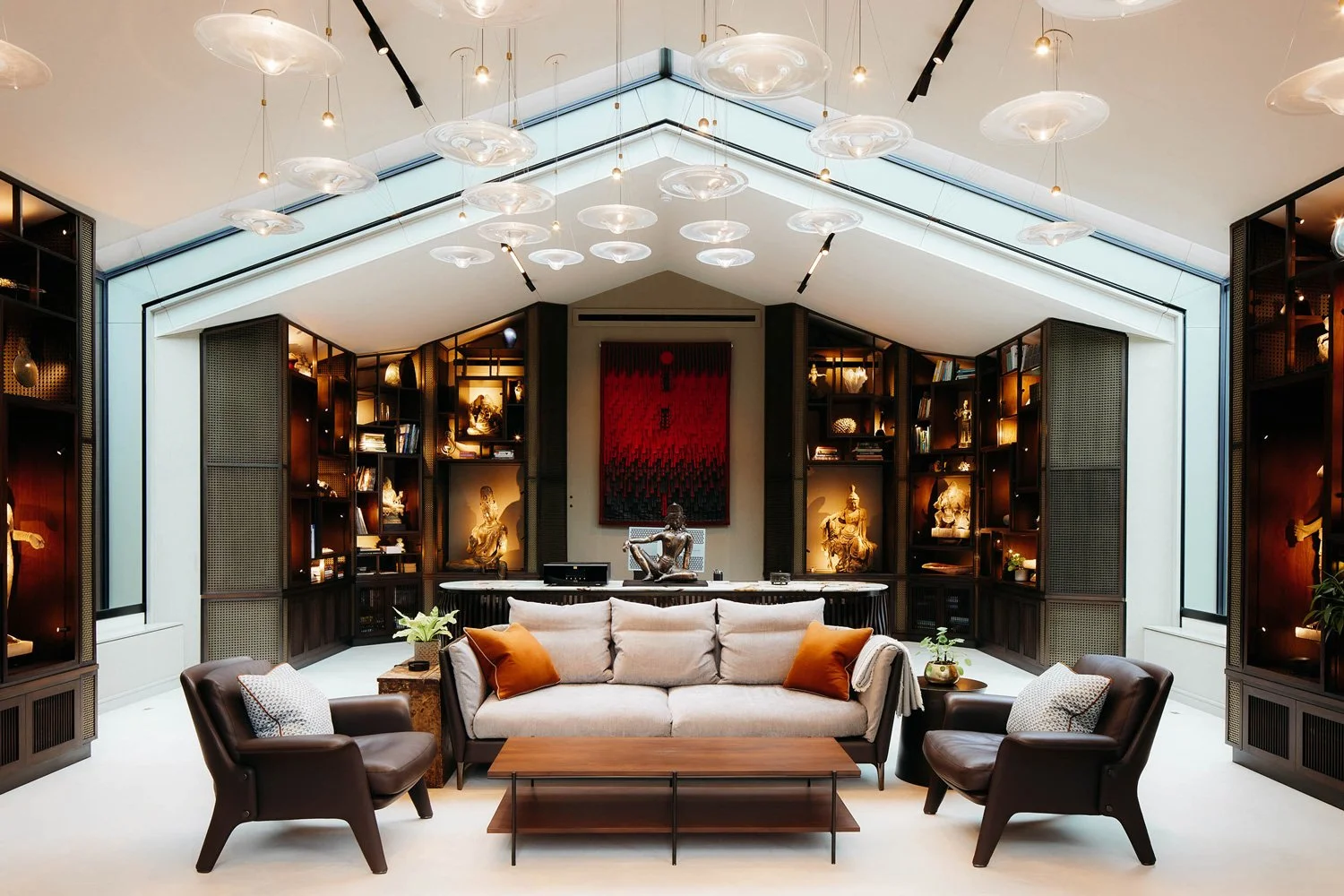 Elegant living room with a beige sofa, two dark leather armchairs, and glass coffee table, surrounded by built-in shelves with sculptures and books, illuminated by modern ceiling lights and natural light from large skylights.