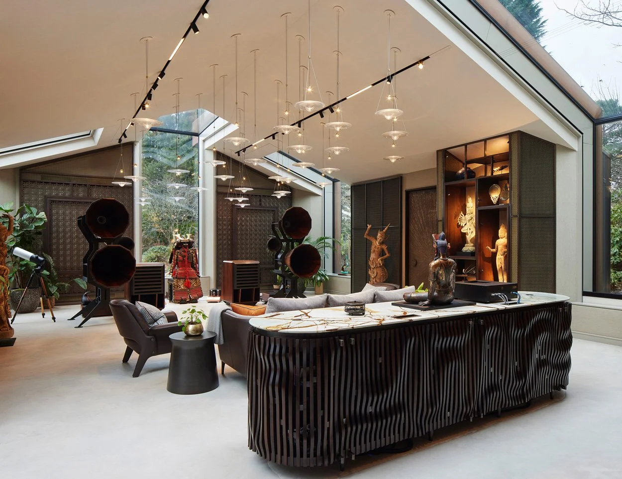 Modern living room with large windows, sculptures, and decorative items, featuring a high ceiling with hanging lights, and a workspace with speakers and a vinyl record player.