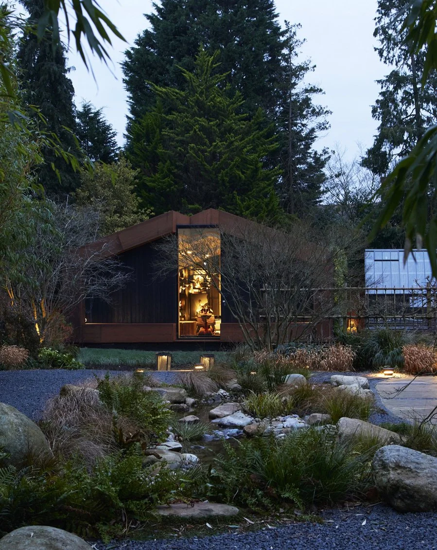 A modern house with large front window illuminated from inside, surrounded by trees and landscaped garden with rocks and small pond, during evening or dusk.