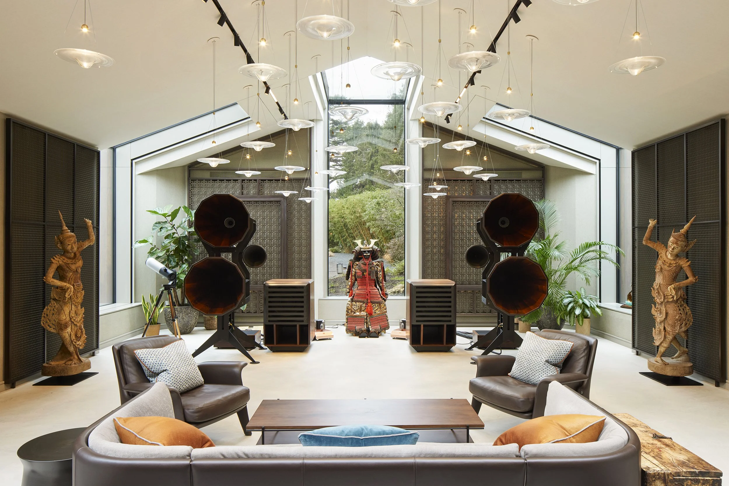 Modern music or listening room with large speakers, leather sofas, decorative statues, plants, and geometric lighting fixtures.