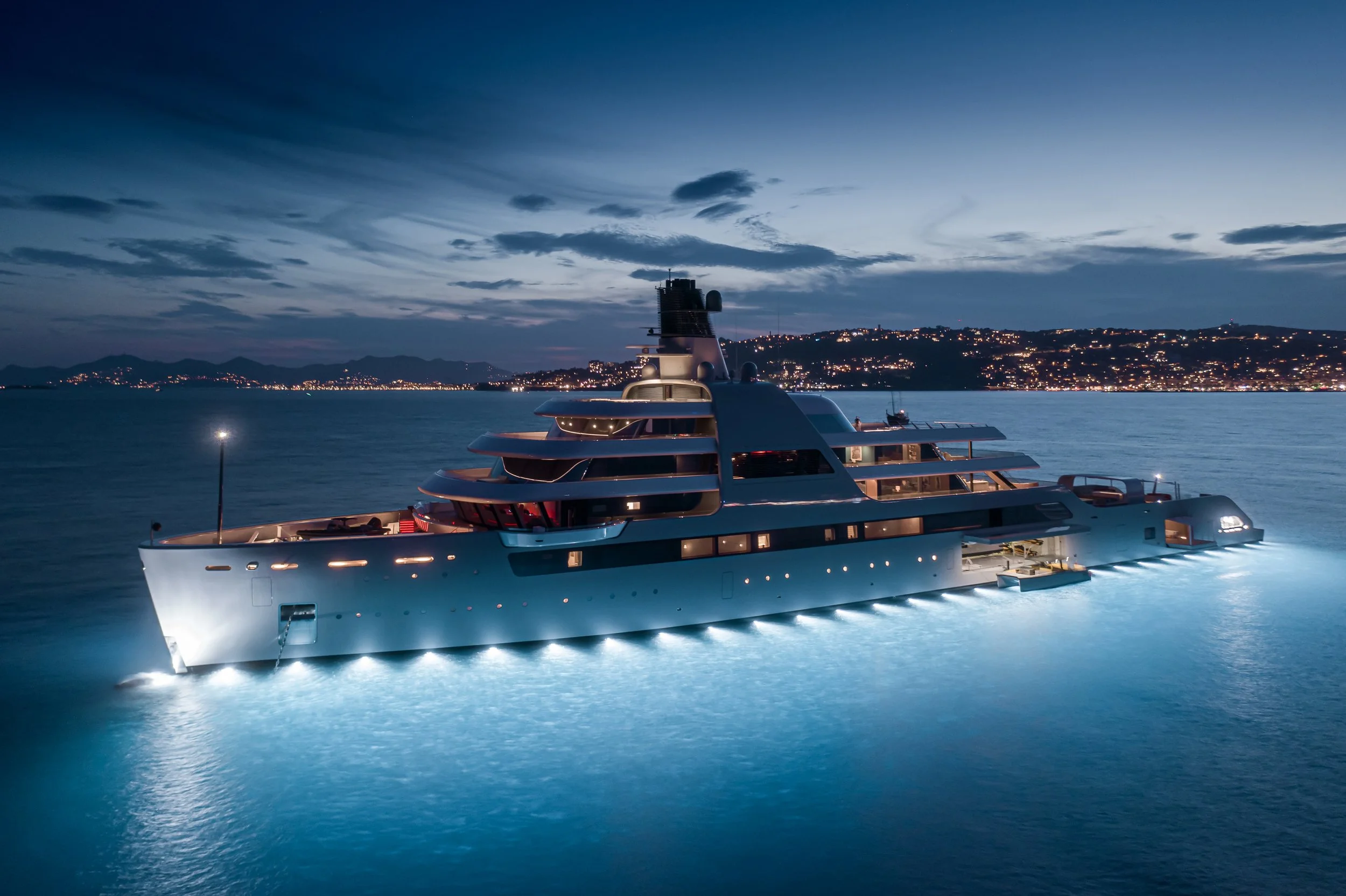 Luxury yacht sailing at night in calm water with city lights on the distant shoreline and a dark sky.