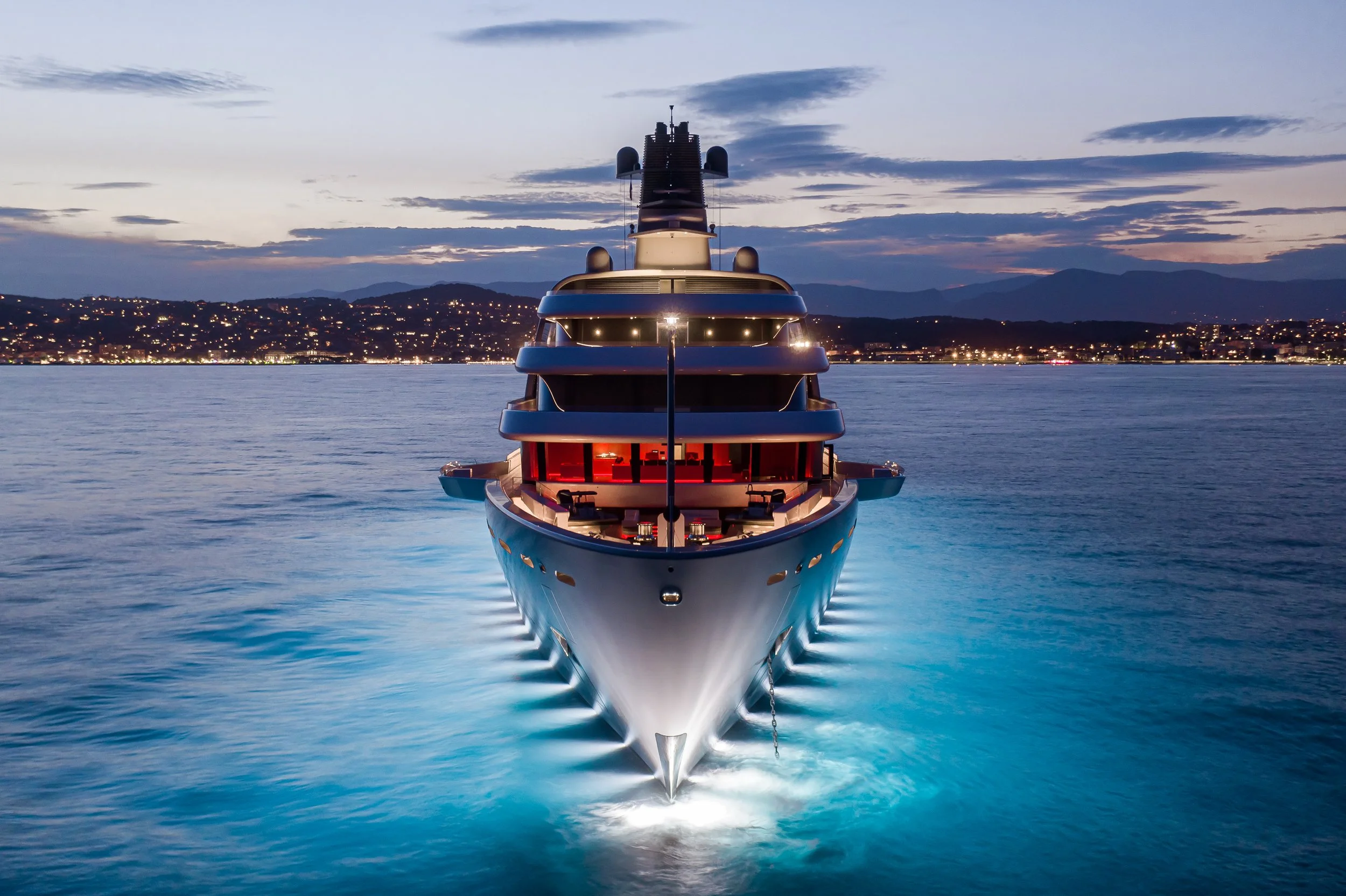 A large luxury yacht sailing on calm water at sunset, with city lights and mountains in the background.