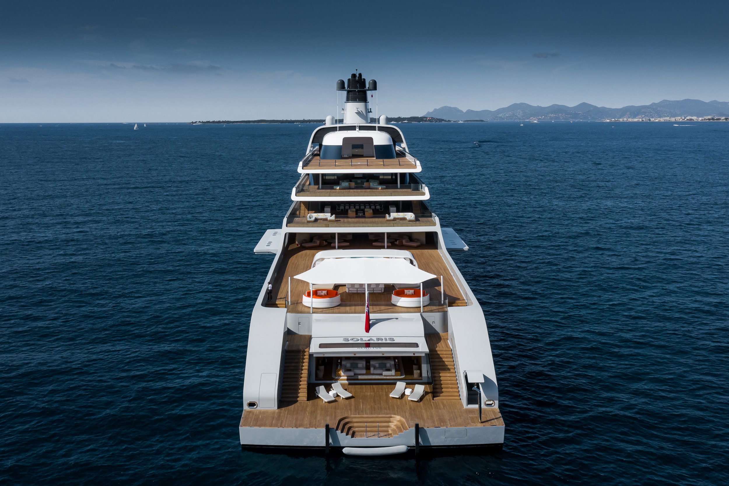 Luxury yacht on open sea with multiple decks and recreational areas, mountains and coastline in background.