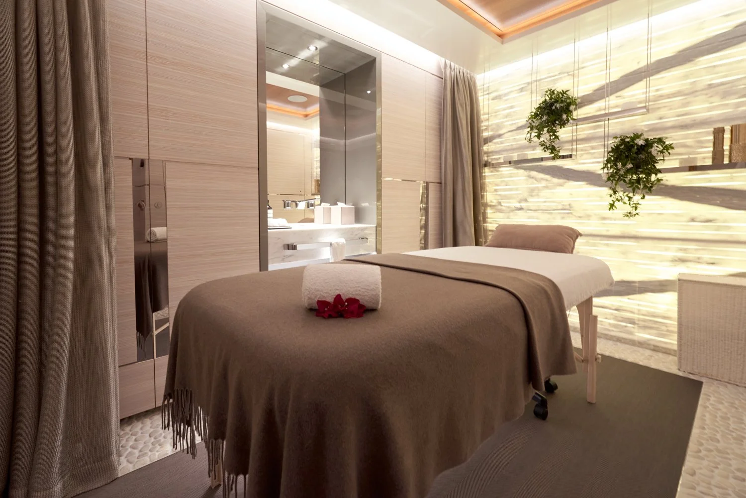 A spa massage room with a massage table covered with a brown blanket, a white towel with red flowers on top, and a pillow. The room has beige walls, curtains, and a textured stone wall with hanging plants. There is a window with sunlight streaming in