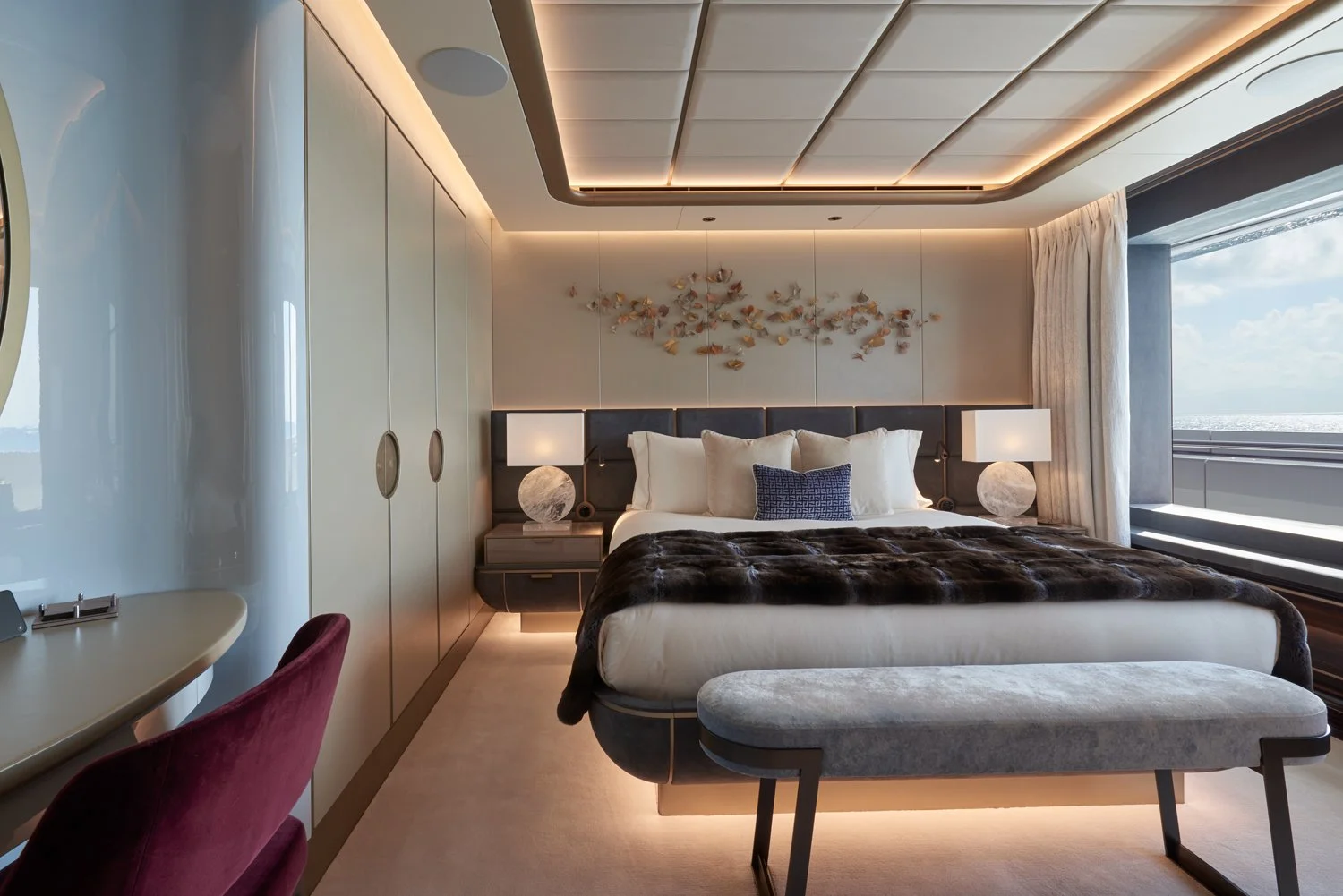 A luxurious hotel room with a large bed, decorative headboard, artwork of birds on the wall, bedside lamps, window view of the ocean, and elegant furniture.