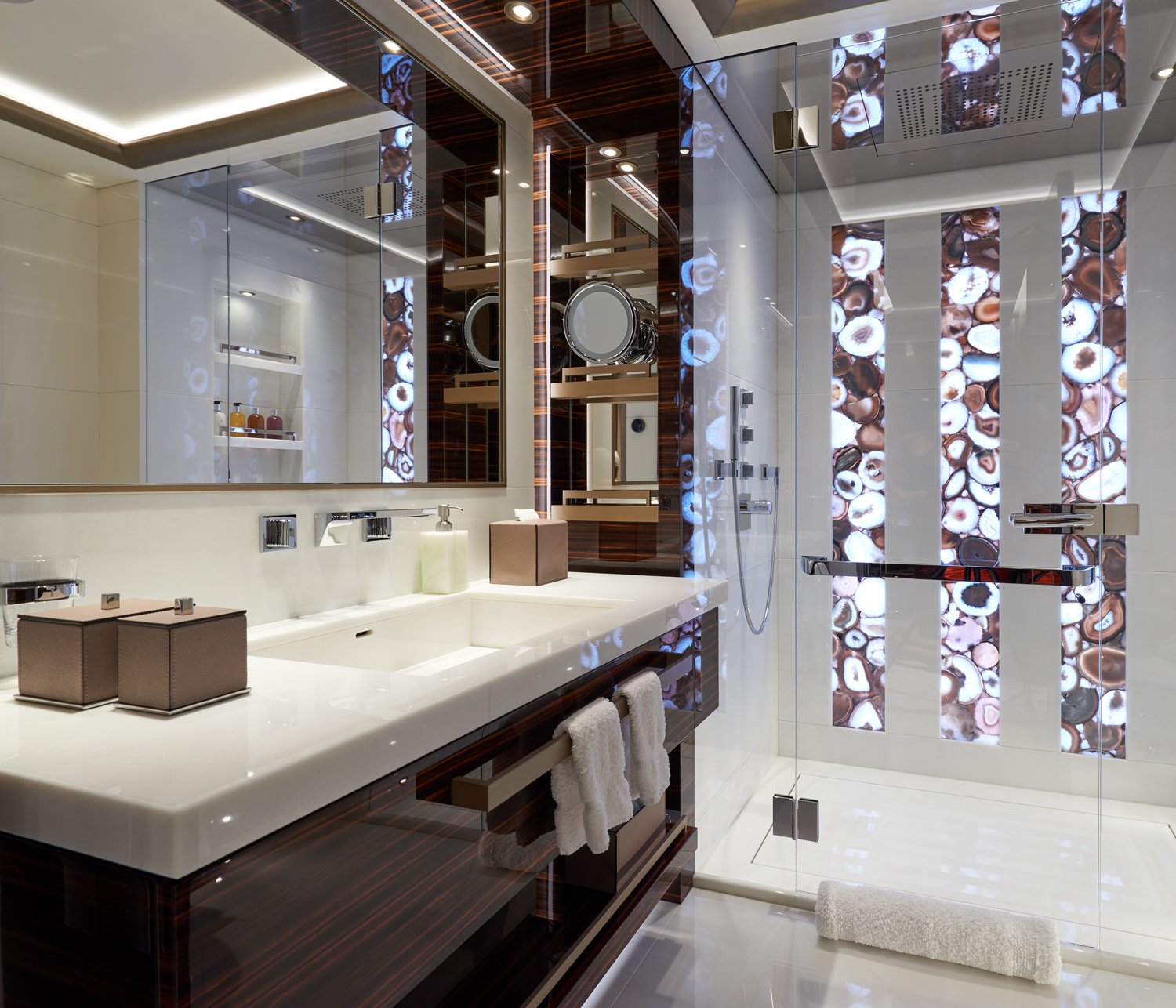 Modern bathroom with a white countertop, large mirror, glass shower with decorative stone pattern, wooden accents, and neatly hung towels.