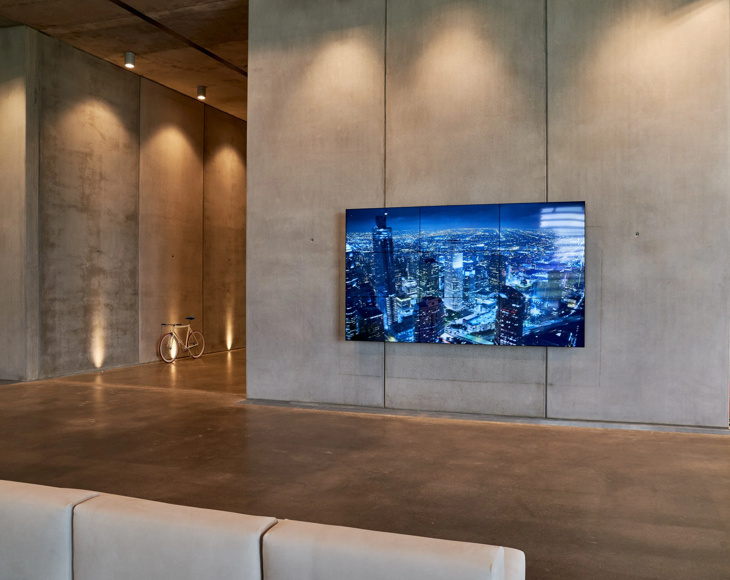 Modern interior with large wall-mounted screen displaying cityscape at night, concrete walls, minimalist lighting, a bicycle, and a beige sofa.
