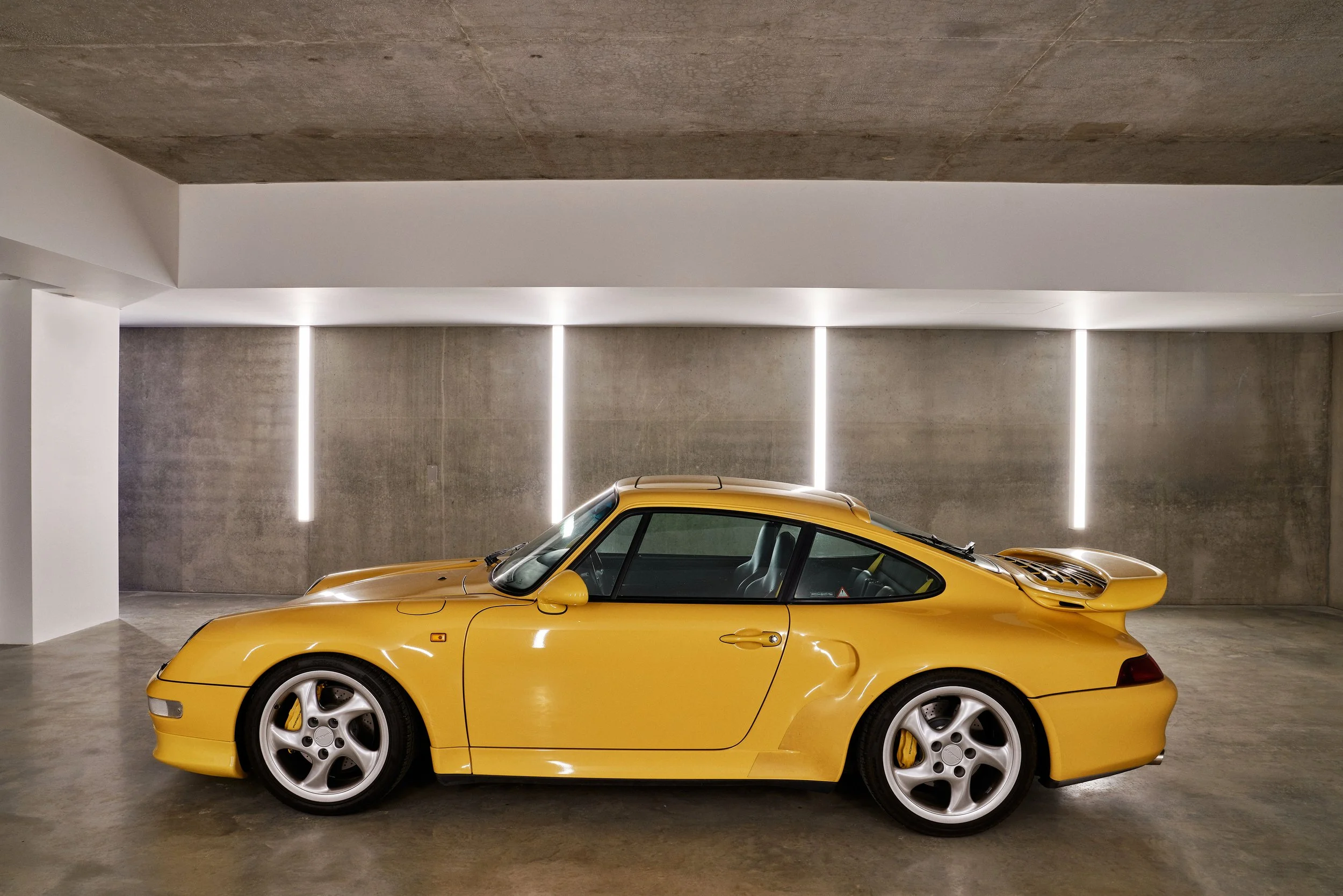 Yellow sports car parked in a modern, minimalistic concrete garage with vertical LED light strips on the wall behind it.