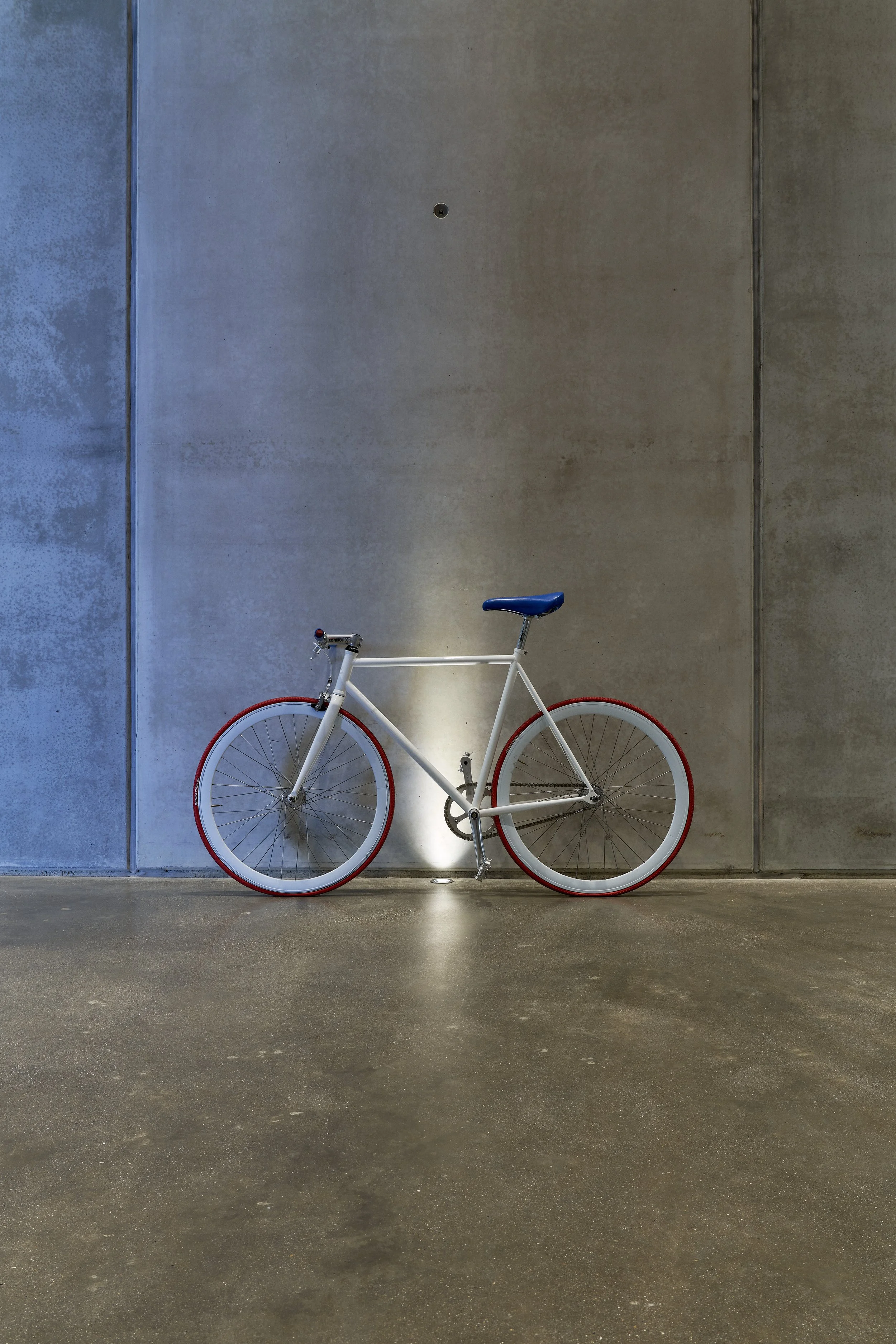 White bicycle with blue seat and red rims against a gray concrete wall in a minimalist setting.