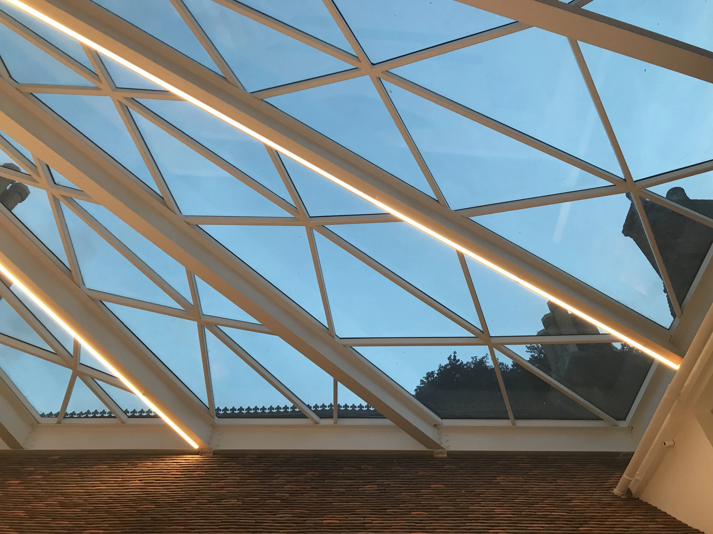 Glass ceiling with geometric metal framework and LED lighting against a blue sky.