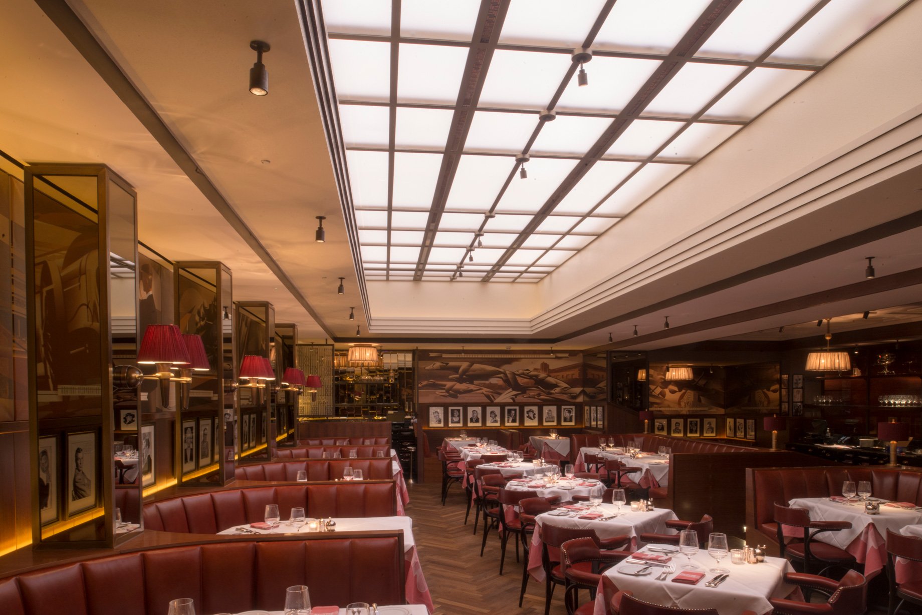 Upscale restaurant dining area with white ceiling skylight, red leather booths, tables set with glasses and utensils, red and beige lamps, and framed black and white portraits on the walls.