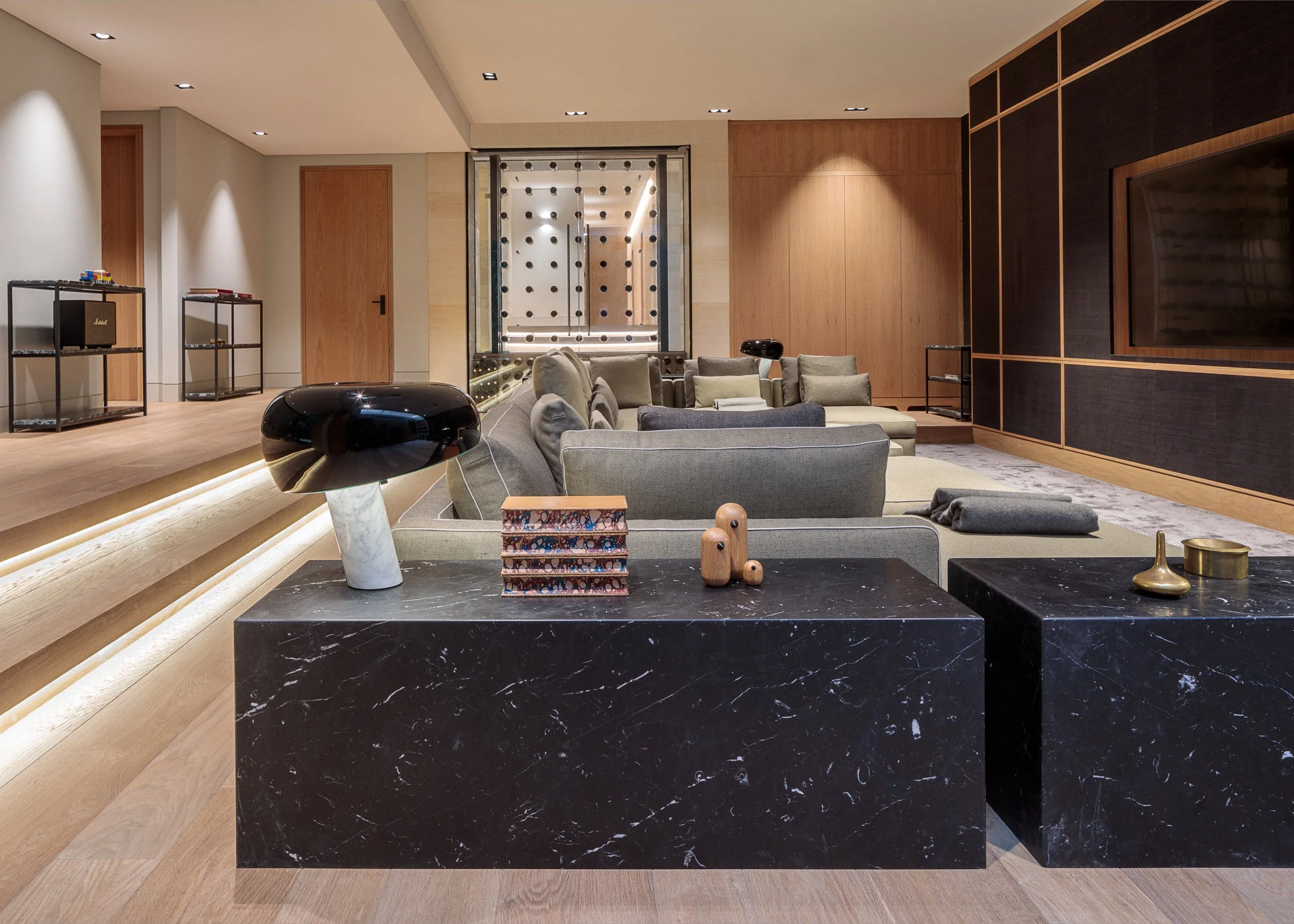 Modern living room with beige and black furniture, decorative items on black marble tables, wooden wall panels, and a large flat-screen TV.