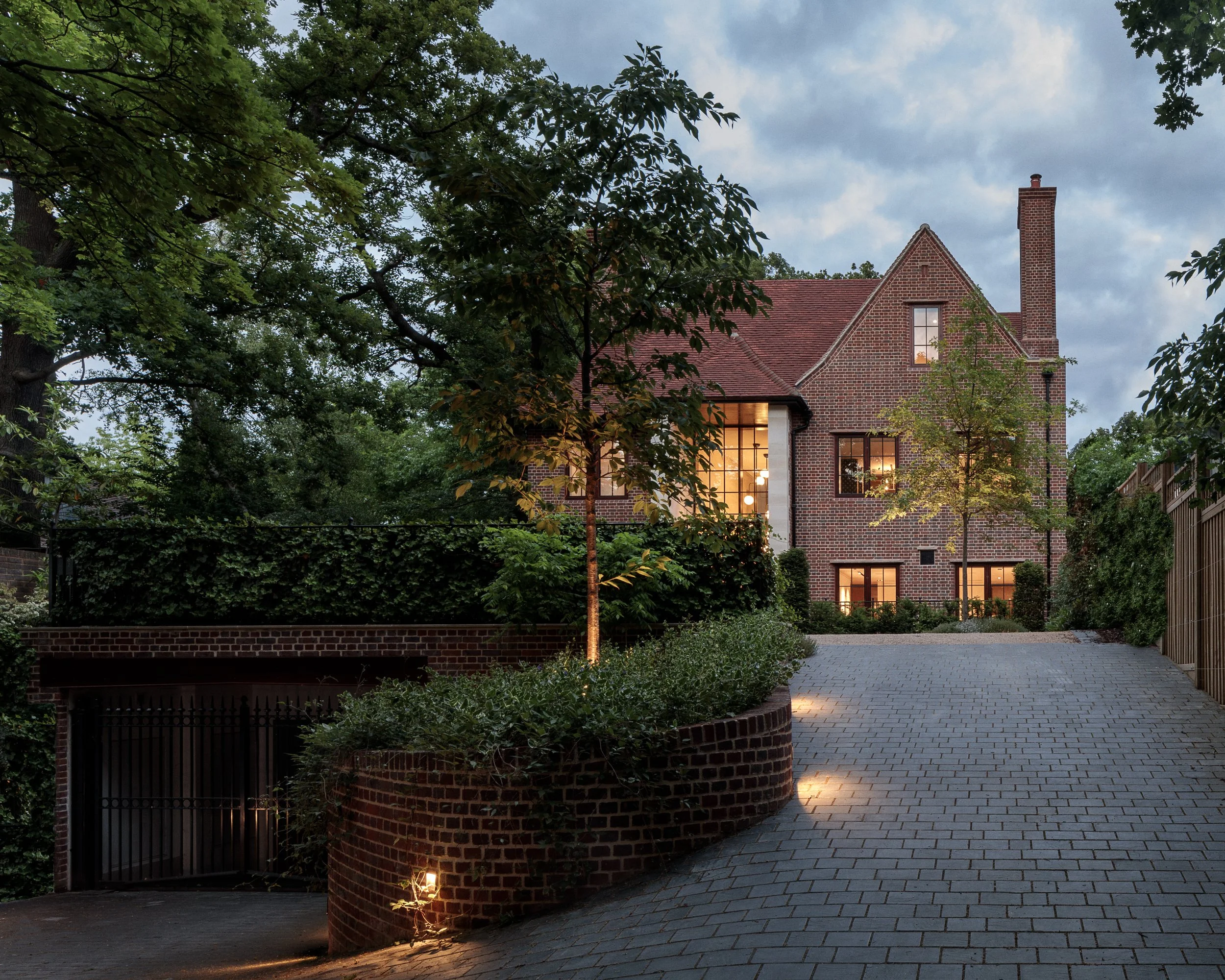 A large brick house with lit windows, surrounded by trees and greenery, with a paved driveway and outdoor lighting at dusk.