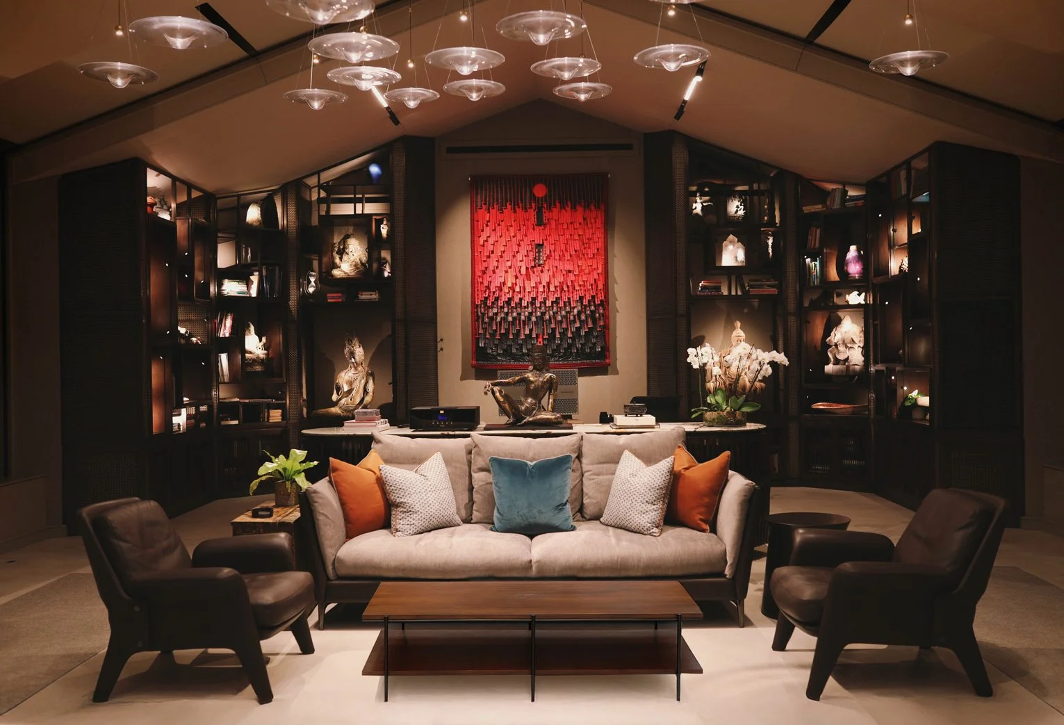 A well-decorated living room with a beige sofa, two black armchairs, and a wooden coffee table. The sofa has throw pillows in orange, white, and blue. Behind the sofa, there is a large bookshelf with decorative sculptures, books, and flowers. An artistic wall hanging with red and black colors is centered on the wall above the sofa. The ceiling has a modern lighting fixture with multiple glass discs.