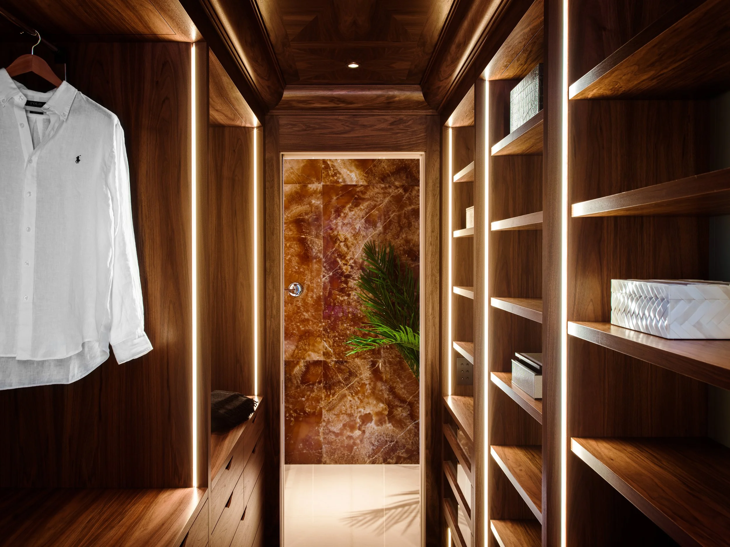 Walk-in closet with wooden shelves and hanging space, illuminated by vertical LED lights, with a marble door and indoor plant at the end.