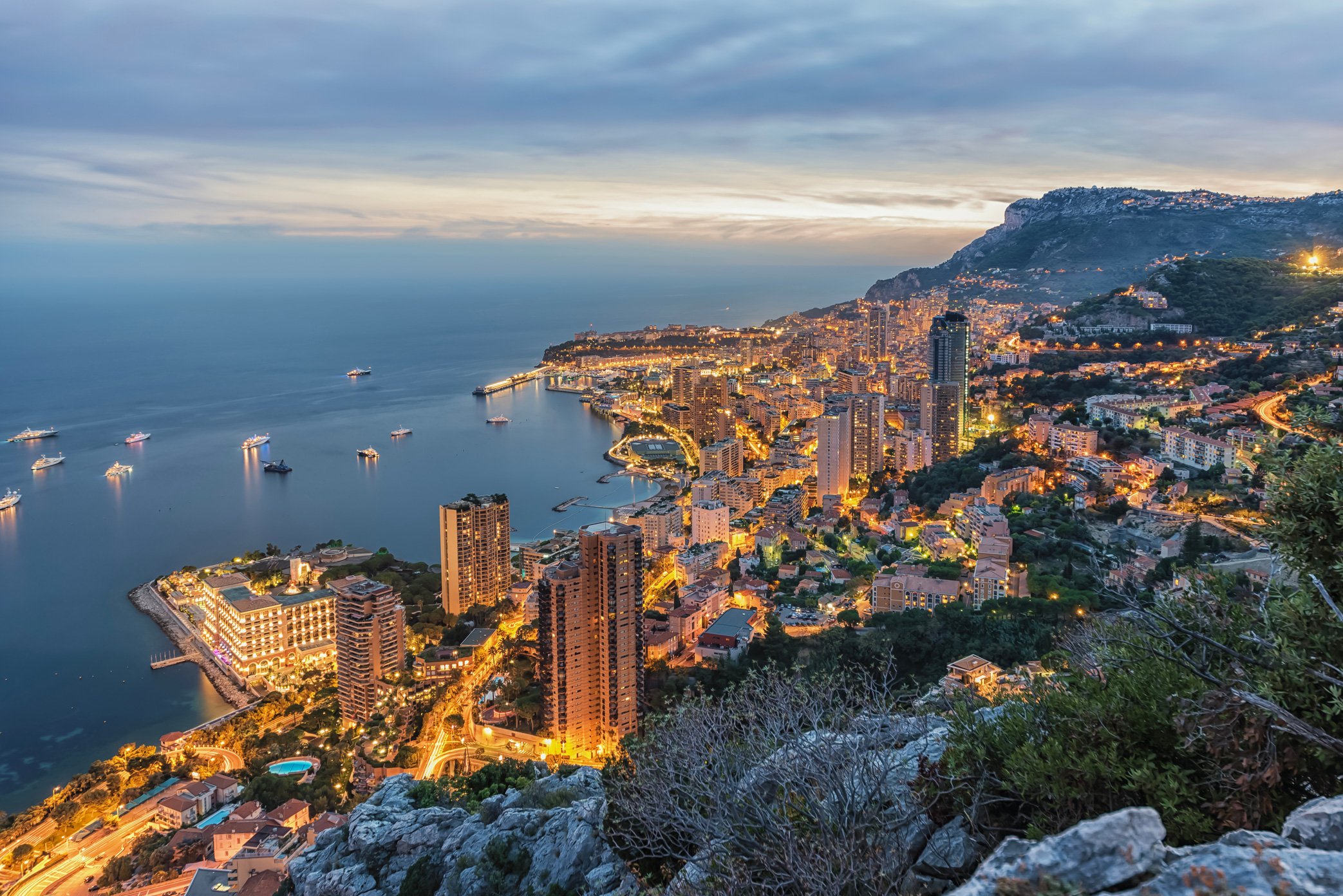 Nighttime view of a coastal city with high-rise buildings illuminated, a marina with boats, and a mountain in the background, seen from an elevated vantage point.