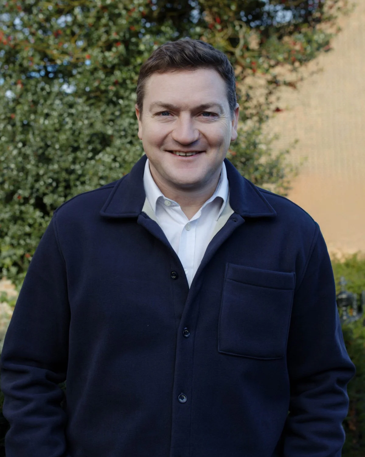 A portrait of a smiling man in a light-colored blazer and white shirt, against a dark blue background.