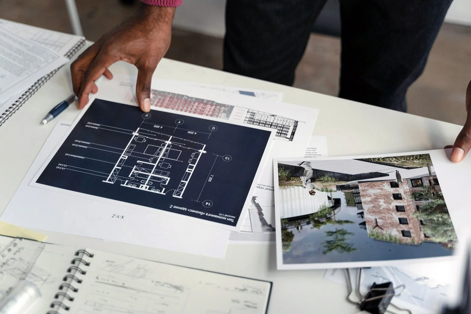 People reviewing architectural plans and design images on a table, including a blueprint and a rendering of a building exterior.