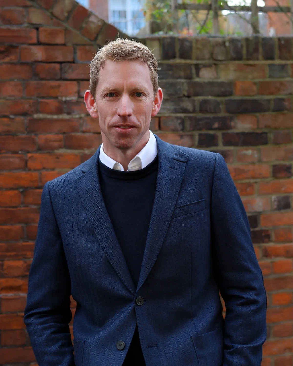 A man with short blonde hair, wearing a blue blazer over a black sweater and white collared shirt, stands outdoors in front of a brick wall.