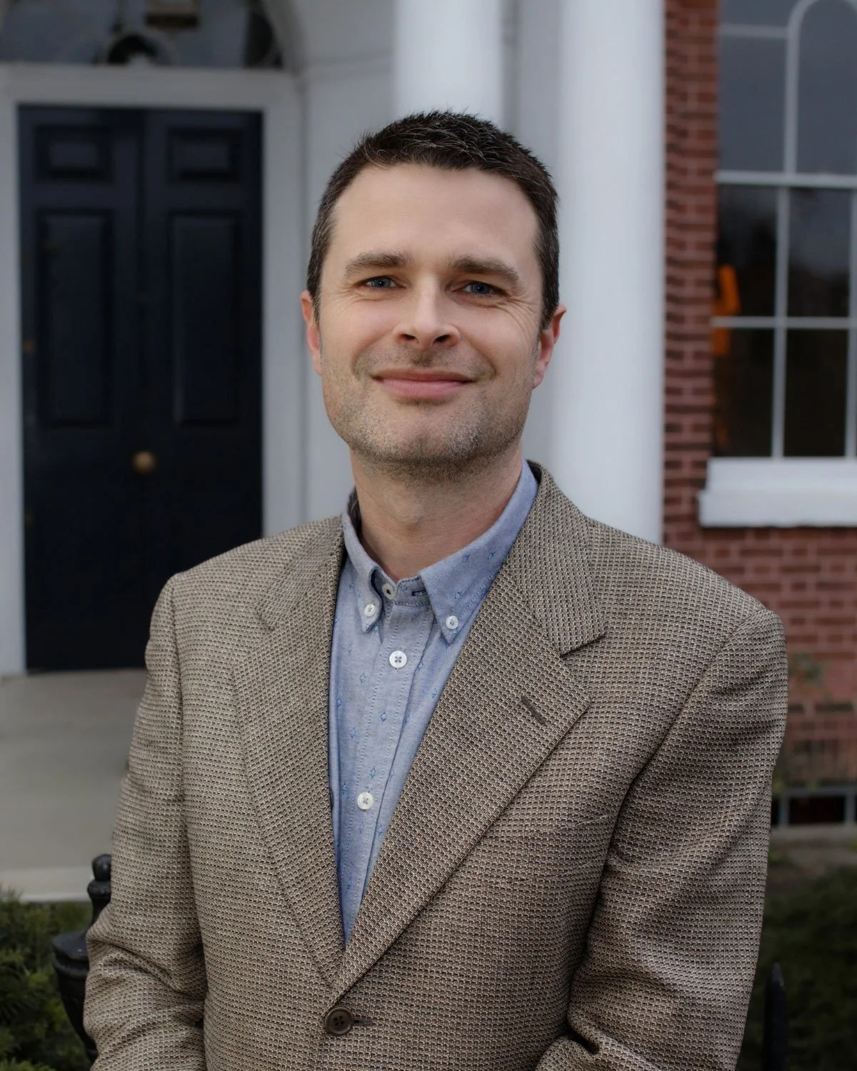 Portrait of a man with dark hair and blue eyes, wearing a white collared shirt, against a blue background.
