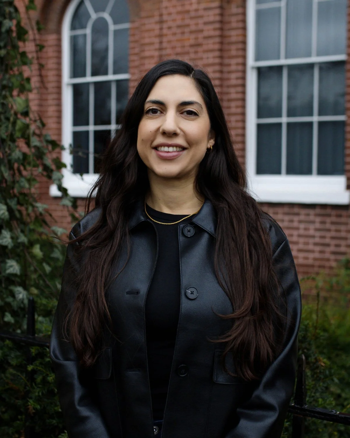 A woman with long brown hair wearing pearl earrings and a black and white striped sleeveless top, standing indoors near a window with green trees outside.