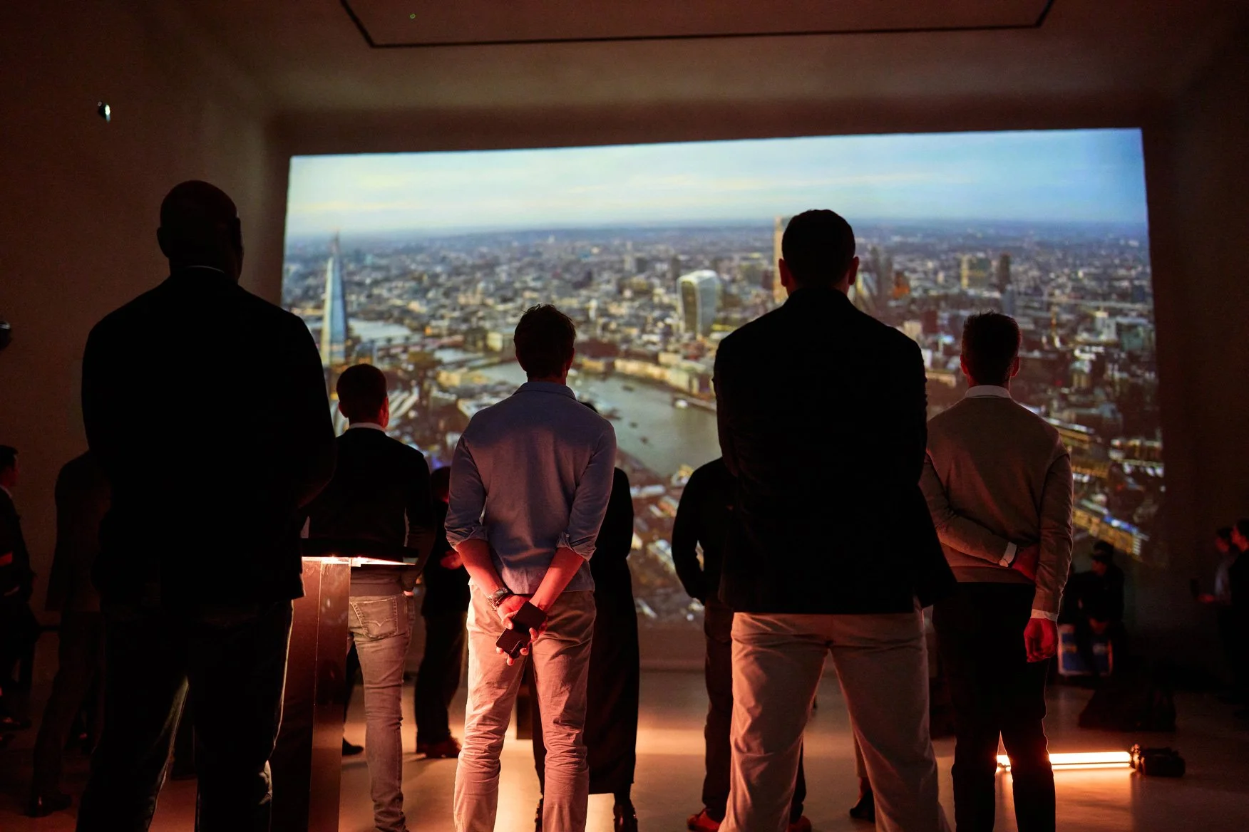 People standing in a dark room watching a large screen displaying a cityscape view of London, including the River Thames and prominent buildings.