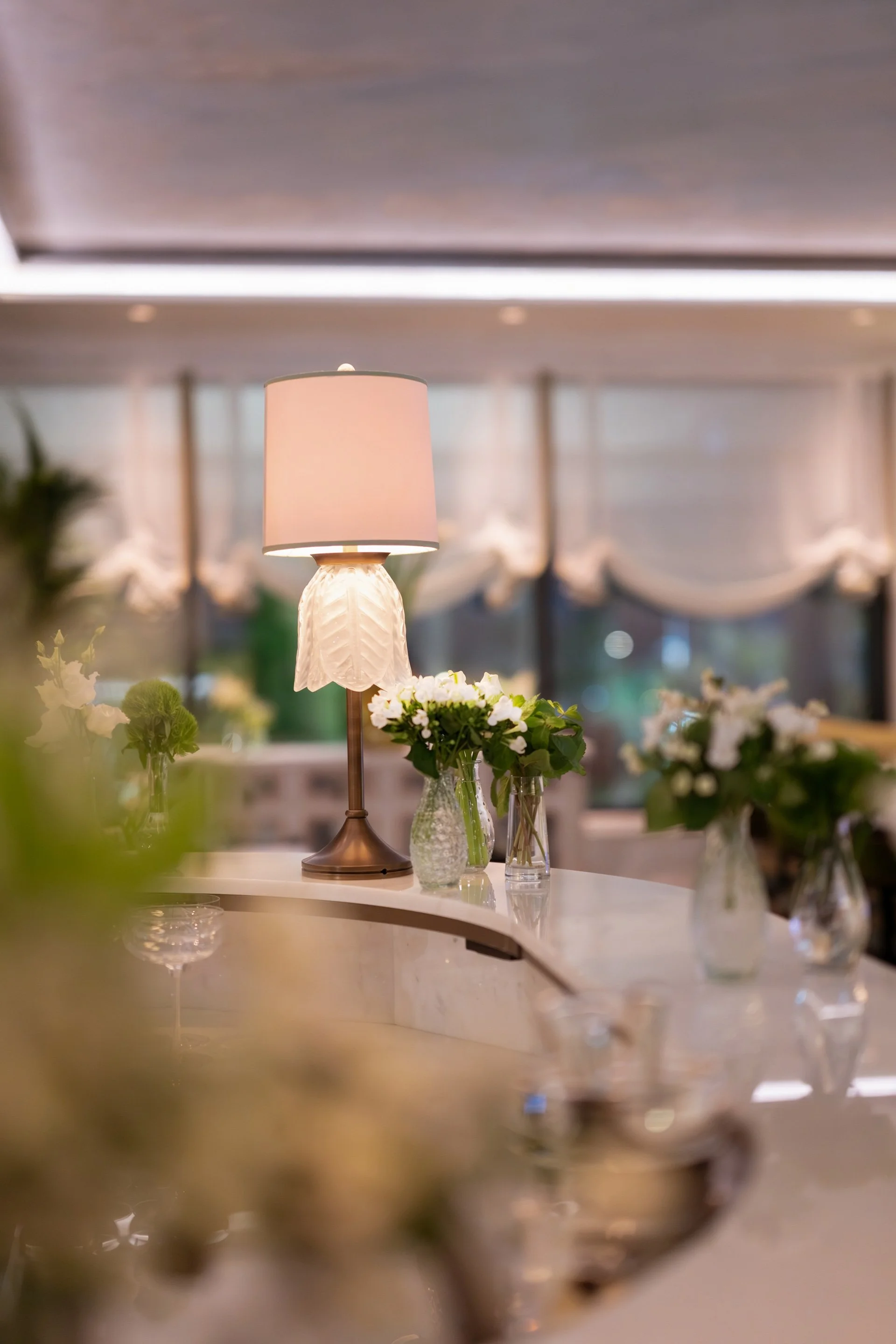 A lit table lamp with a white shade and decorative base, surrounded by glass vases with white flowers on a marble countertop in an elegant, softly lit room.