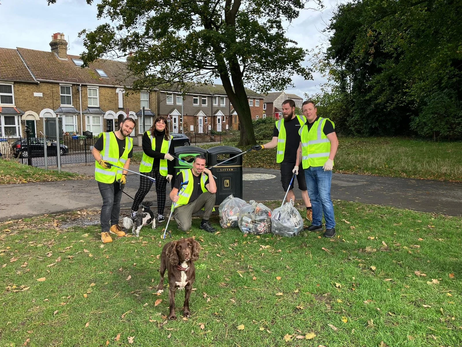 A group of six volunteers, five men and one woman, wearing yellow safety vests and gloves, picking up litter in a park with their dogs. They are standing near a trash bin filled with collected trash, smiling for the camera.