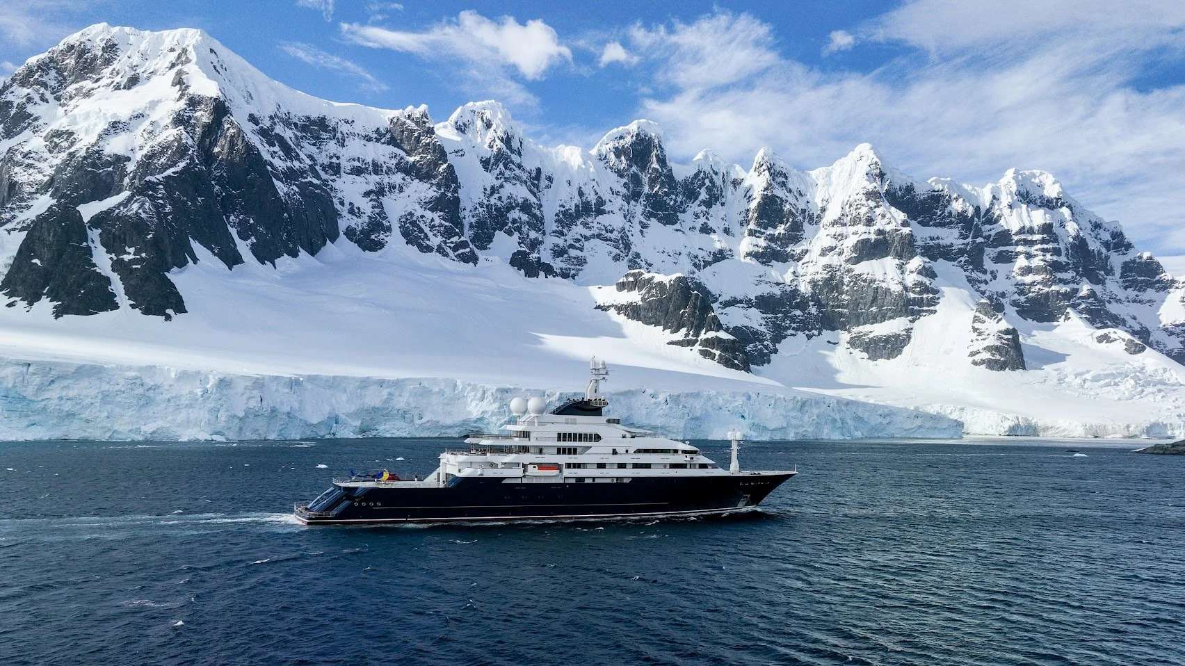 Luxury yacht sailing near icy Antarctic landscape with snow-covered mountains and glacier in the background.
