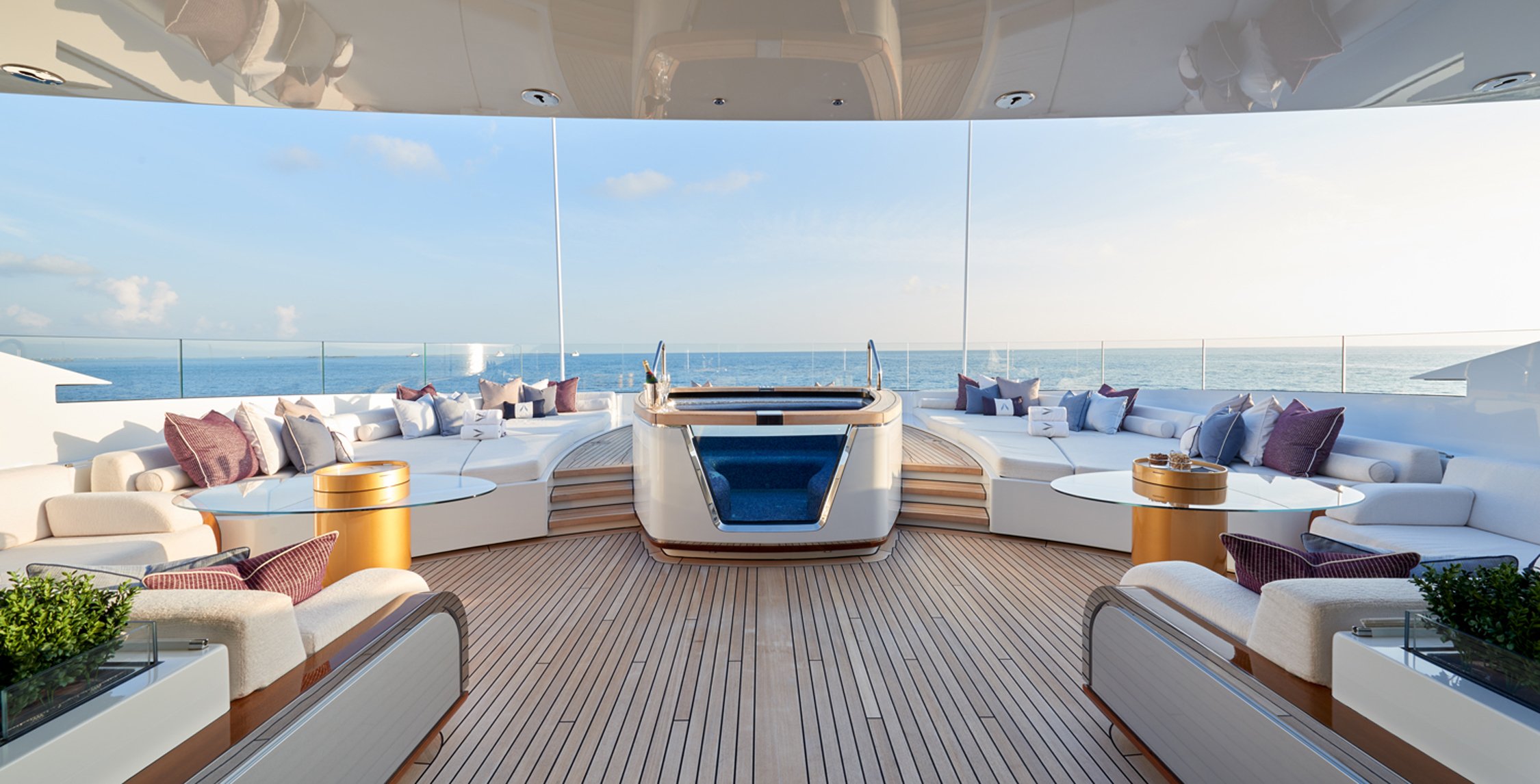 Luxury yacht deck with white cushioned seating, pillows, glass tables, a hot tub, overlooking the ocean under a clear sky.