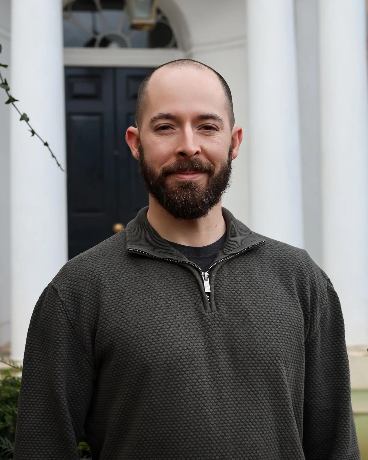 A man with a beard and a shaved head standing in front of a white building with a dark door and white columns.