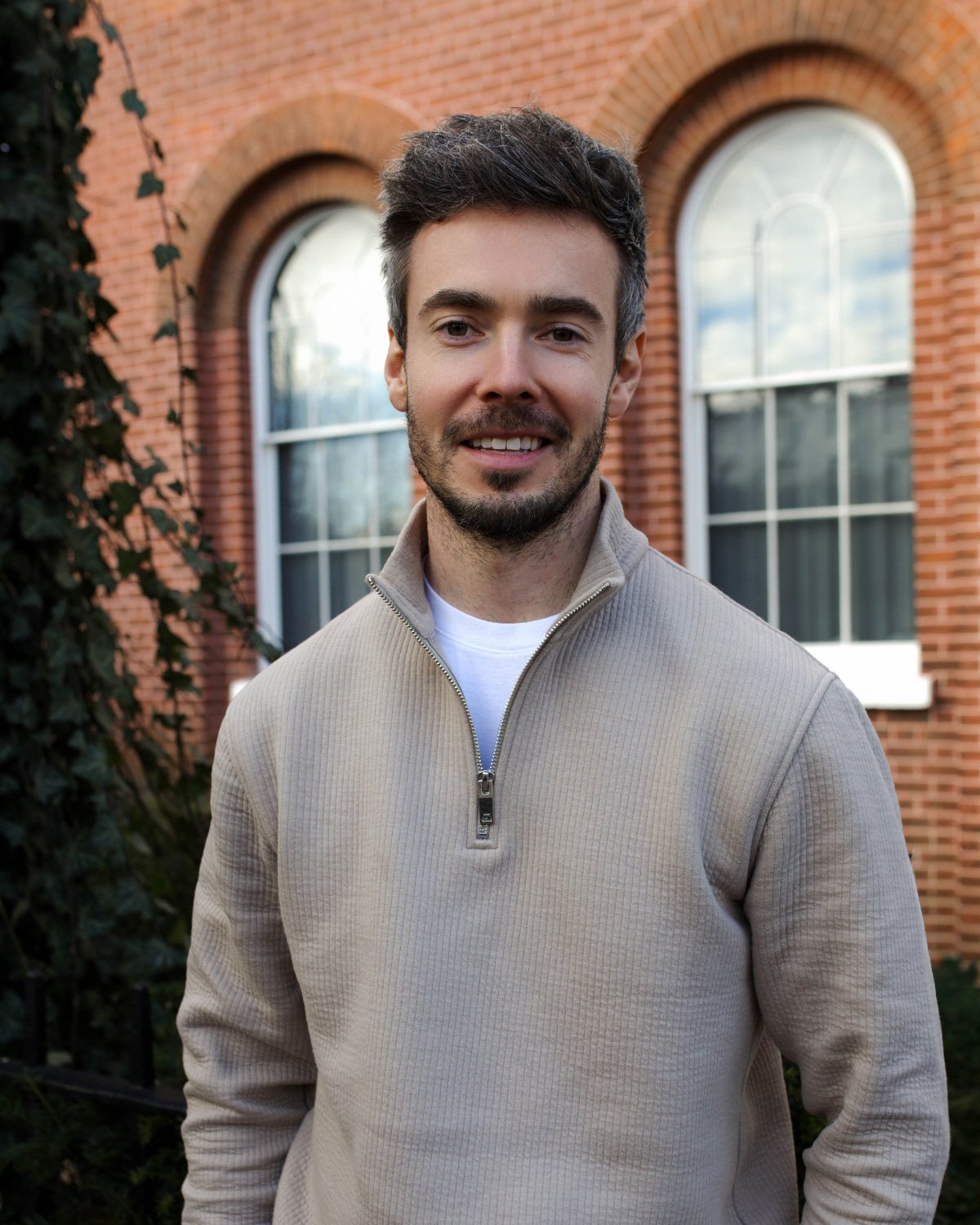 A young man with light skin, dark hair, and a short beard, wearing a light gray button-up shirt with a Polo Ralph Lauren logo, posing against a blue gradient background.