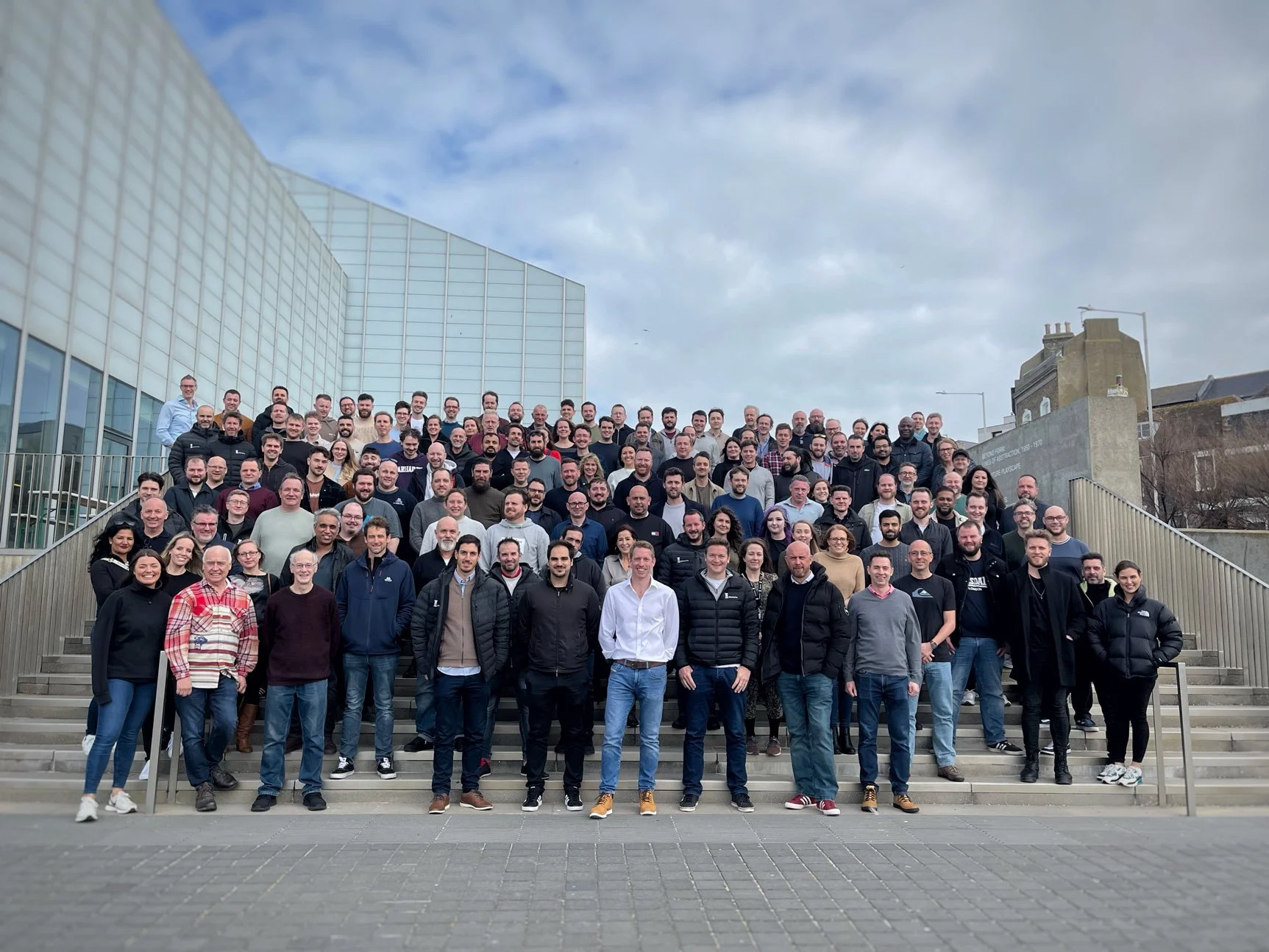 A large group of people posing on outdoor steps, with a modern glass building on the left and older buildings on the right, under a cloudy sky.